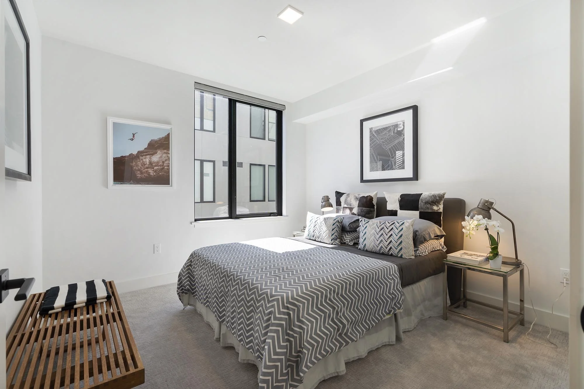Modern bedroom with white walls, a window, and black-and-white artwork. A bed with a gray and white zigzag patterned duvet and matching pillows is along the wall. A nightstand with a flower vase, books, and a lamp is beside the bed. A wooden bench is
