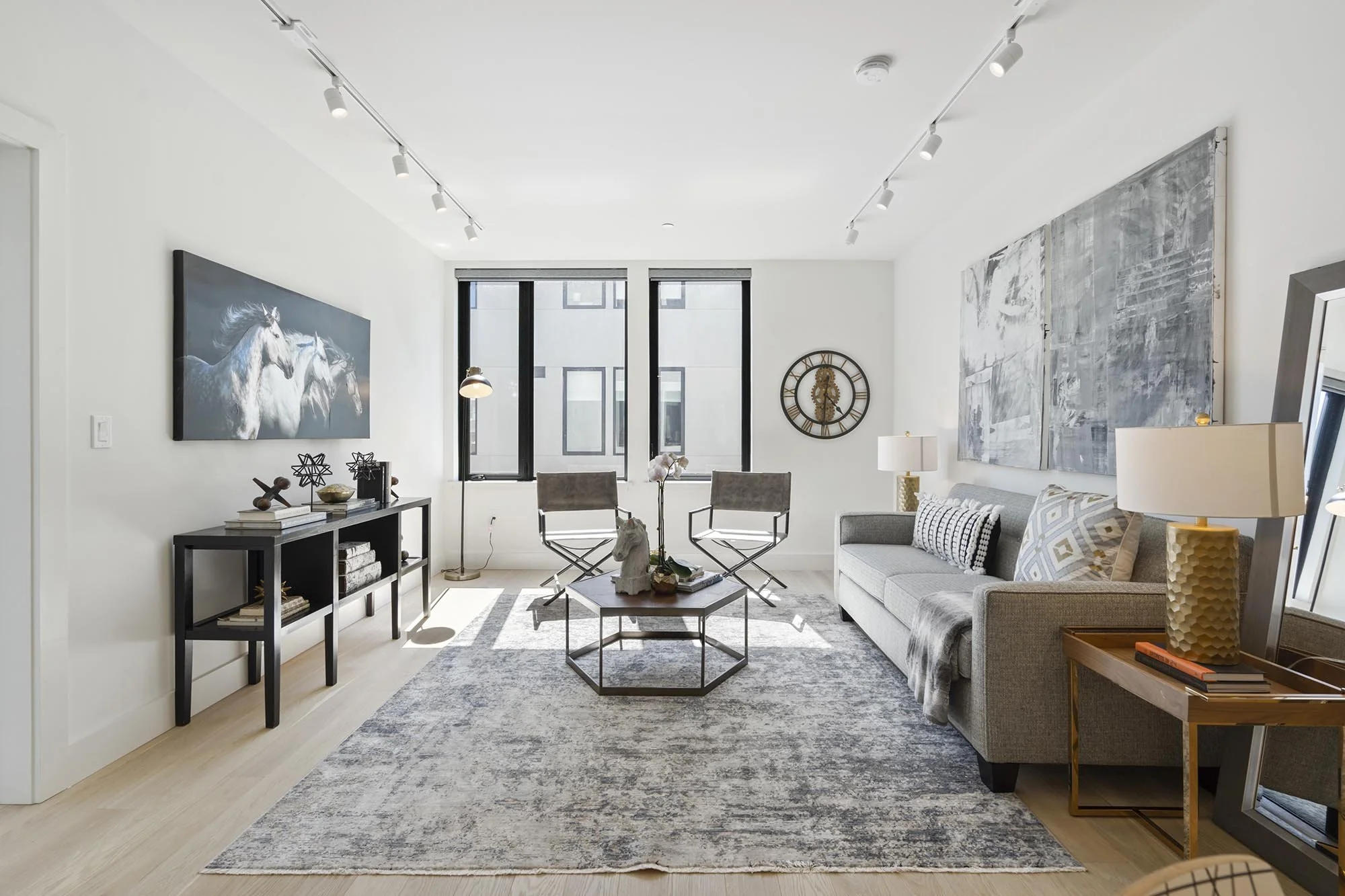 Modern living room with white walls, large windows, gray sofa, framed artwork, decorative items on a black console, and two chairs, on a gray patterned rug.
