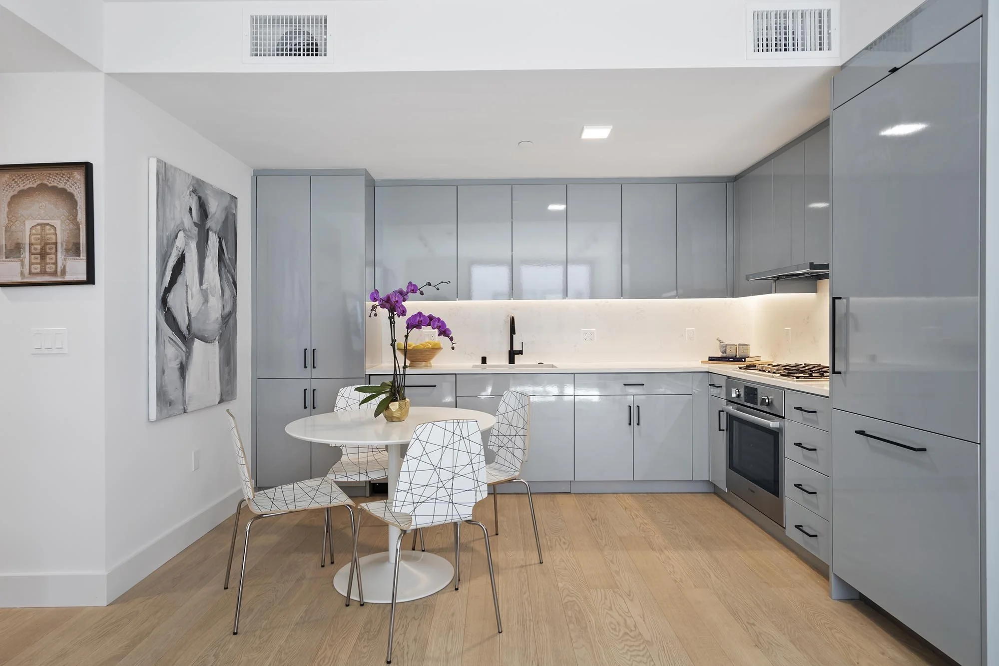 Modern kitchen with light gray cabinets, white countertops, and a small round white dining table with four wire-frame chairs. A purple orchid plant is on the table. There are two abstract black-and-white paintings on the wall.