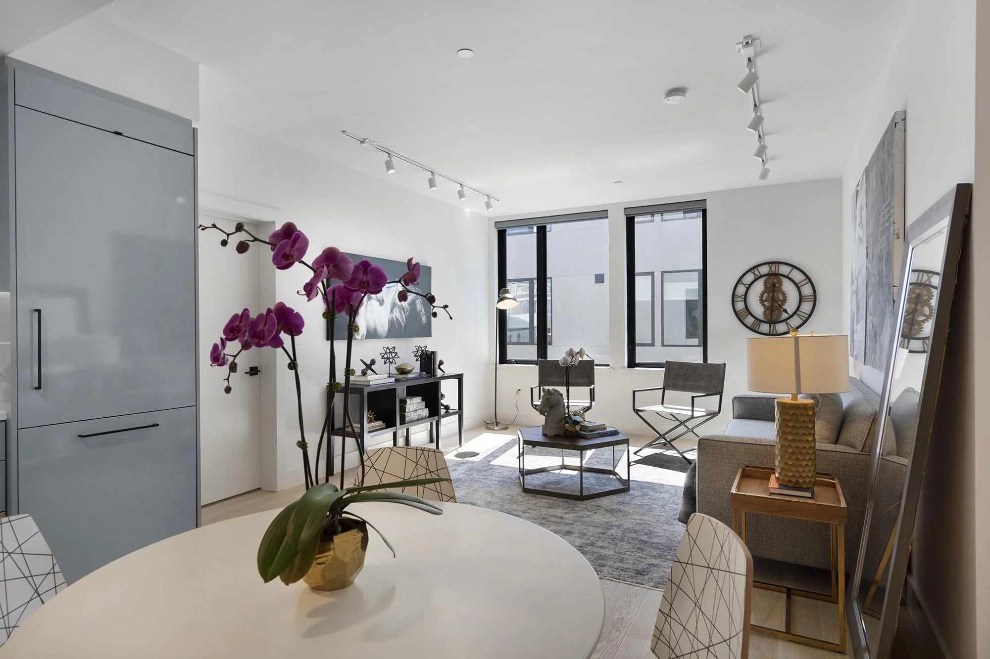 Modern living room with large windows, gray sofa, chairs, a clock on the wall, plants, and artwork, illuminated by natural light and ceiling track lighting.