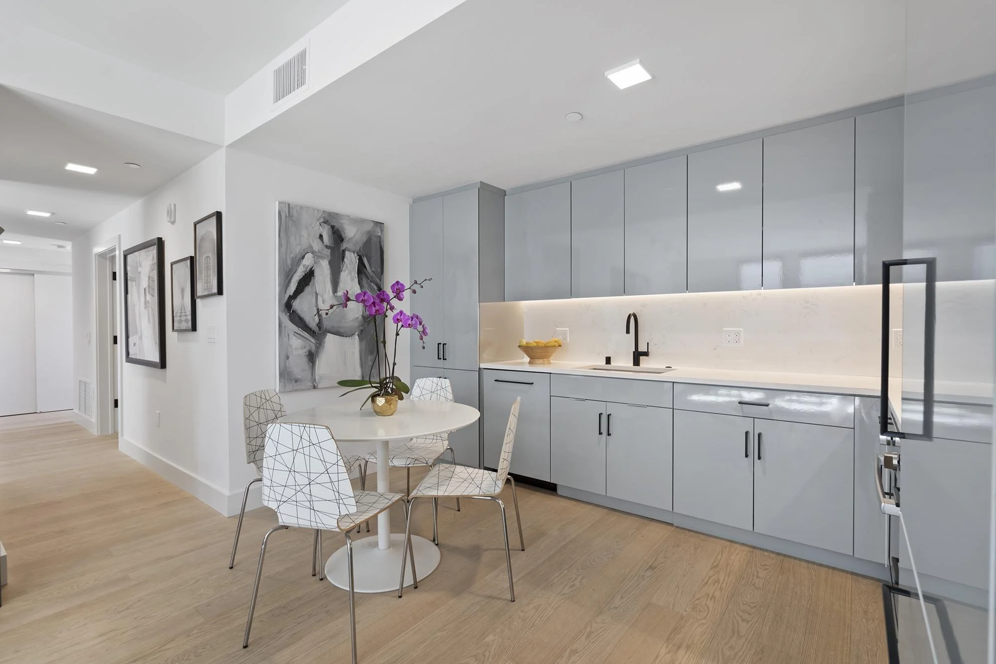 Modern kitchen with light blue cabinets, white marble backsplash, black sink faucet, round white dining table with white wireframe chairs, a purple orchid in a gold pot, and framed black and white artwork on the wall.