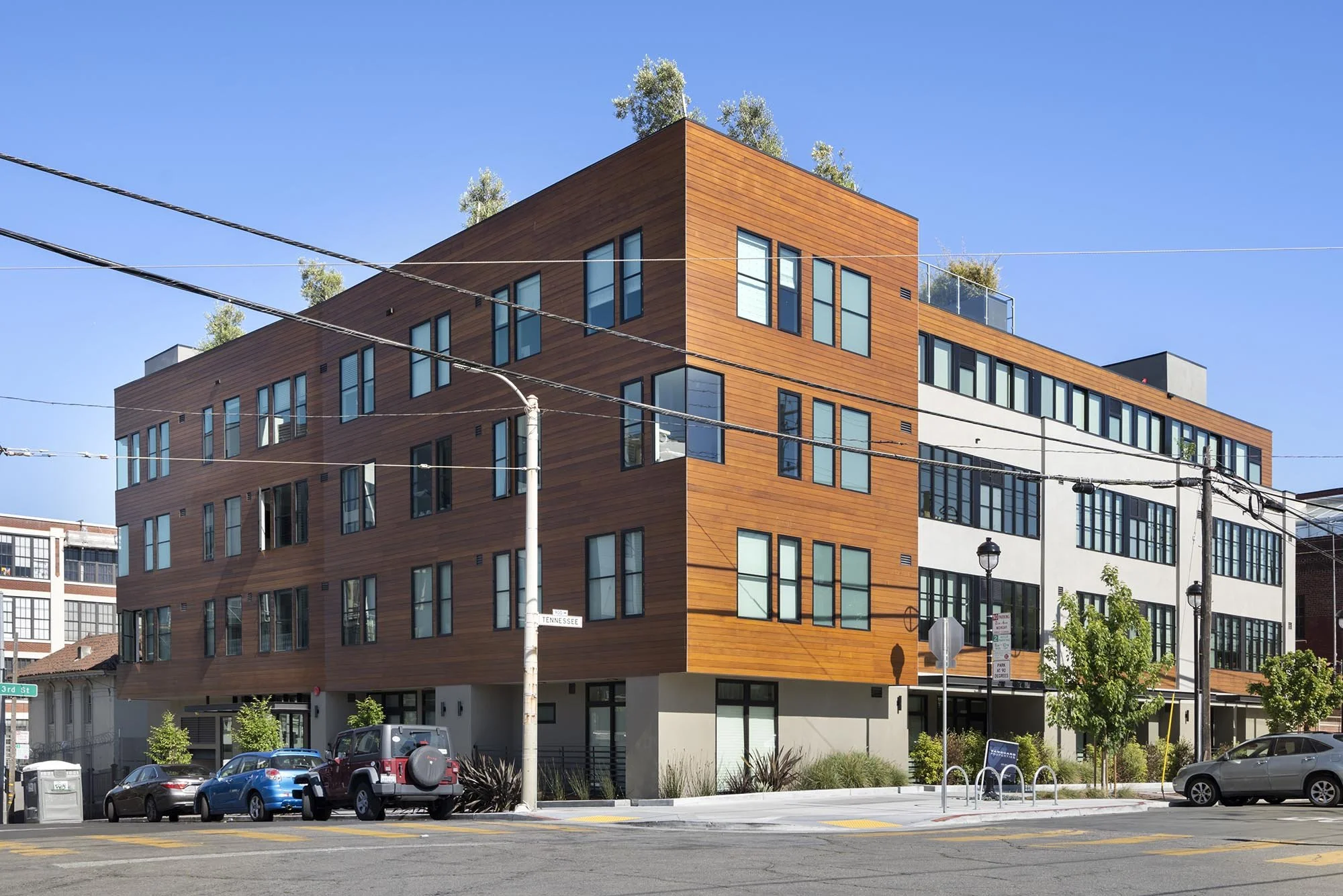 Modern multi-story building with wooden facade and many windows, situated at a street corner under a clear blue sky, with cars parked in front and trees on the roof.