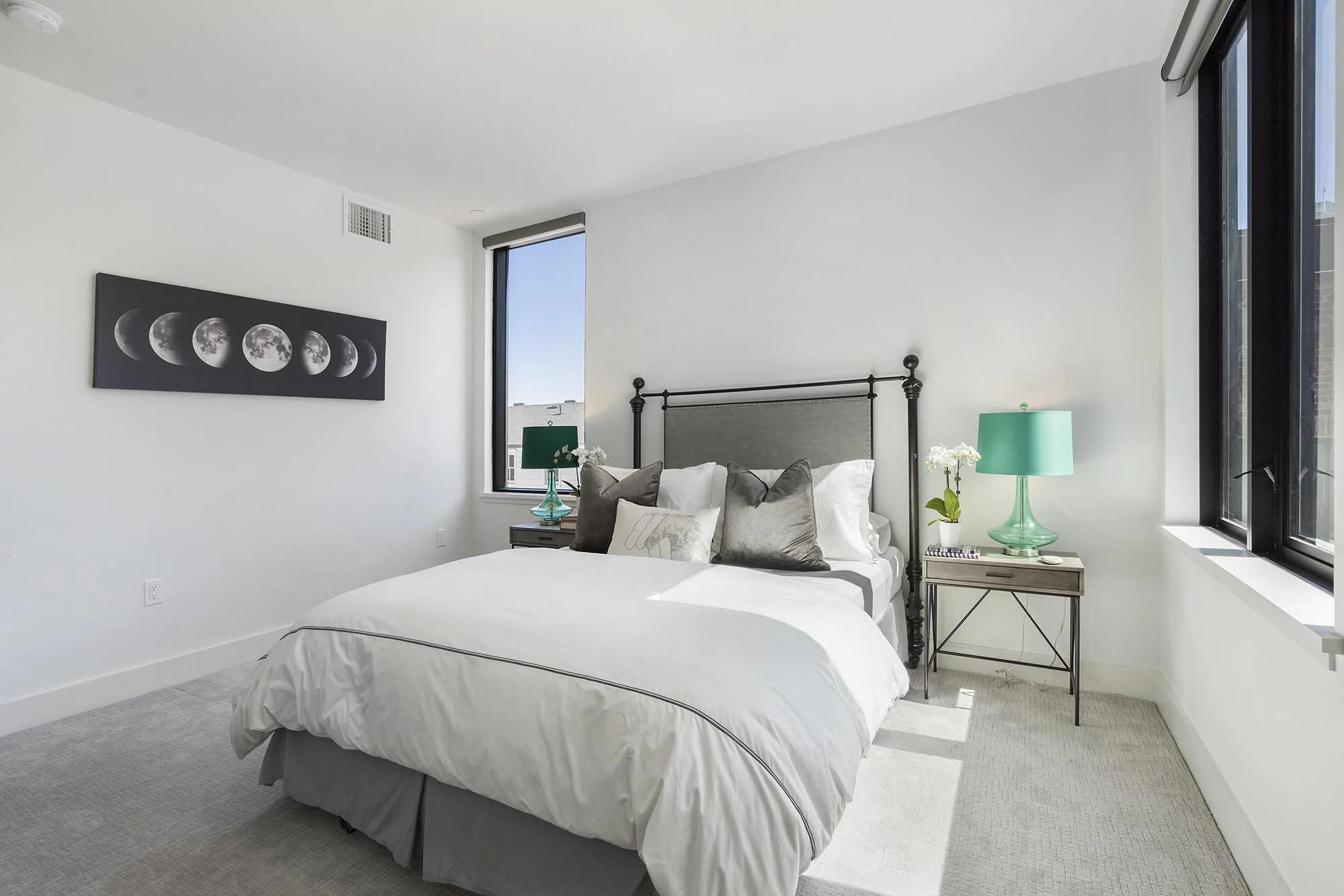 A modern bedroom with white walls, a bed with white bedding, dark pillows, and a metal headboard. Two windows let in natural light, and on one side is a nightstand with a turquoise lamp, a small plant, and accessories. A moon phase artwork hangs on t