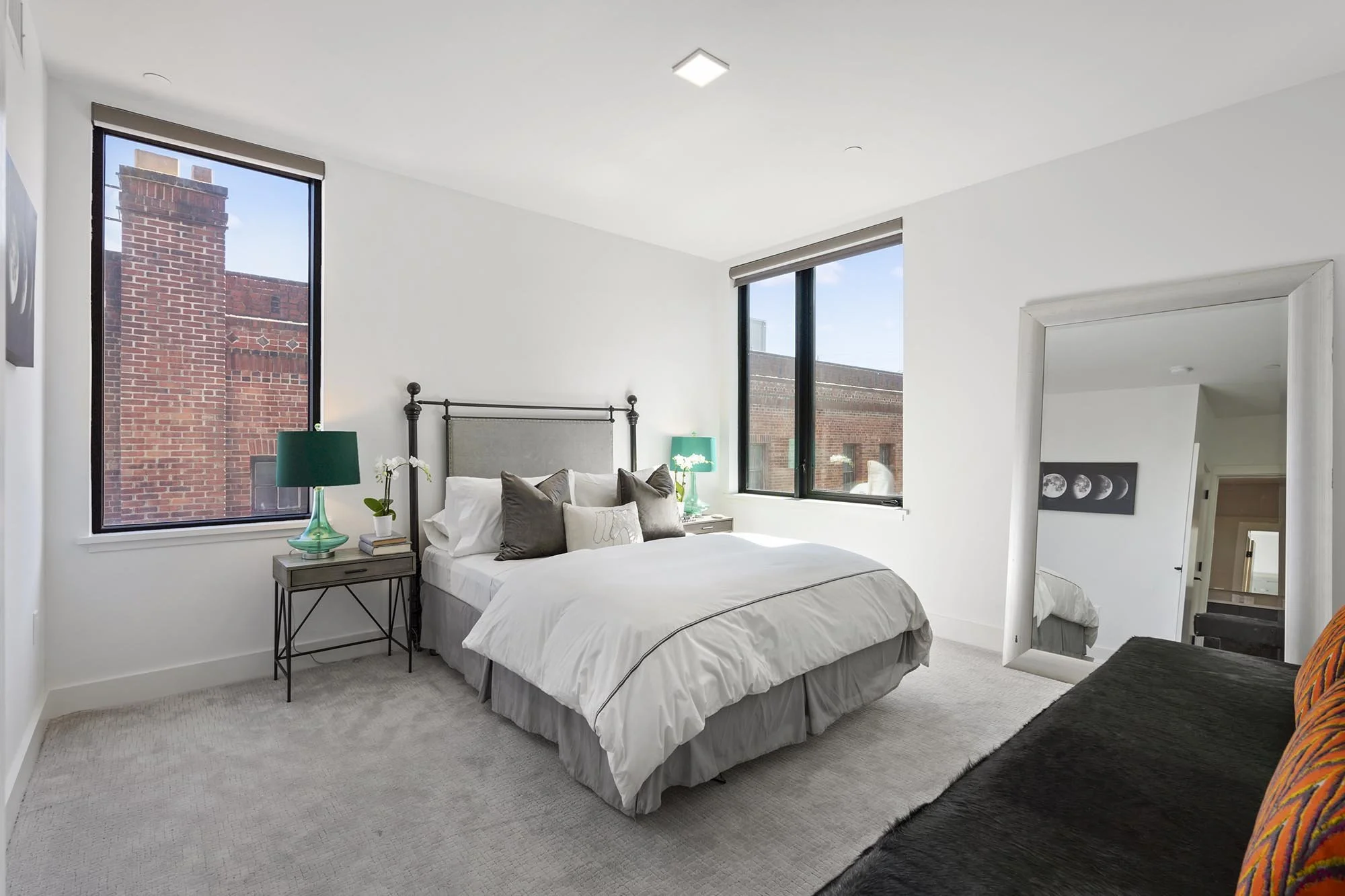 A bedroom with white walls and large windows showing a brick building outside. There is a bed with white and gray bedding, a black metal headboard, and several pillows. On each side of the bed, there are teal lamps on bedside tables. A standing mirro
