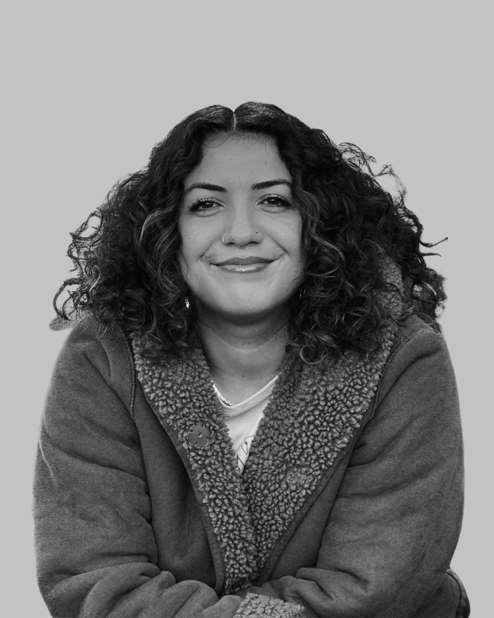 A woman with long curly hair smiling, wearing a jacket with shearling collar, sitting with her arms crossed in front of her. The picture is in black and white.