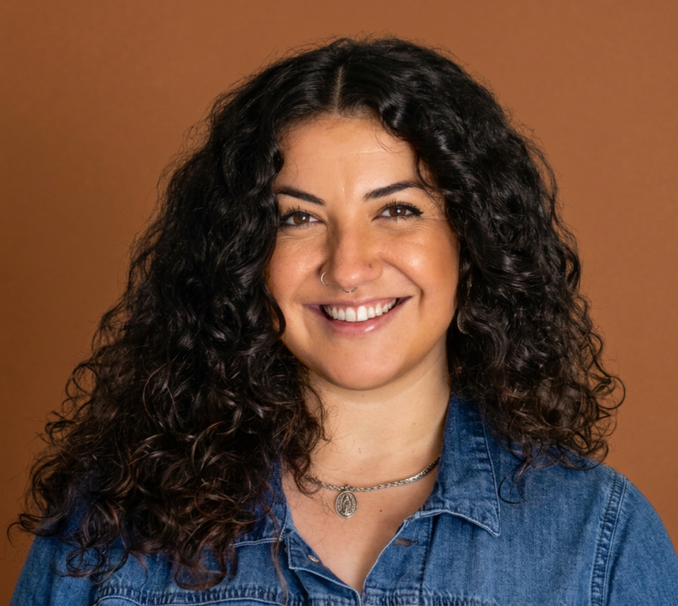 Portrait of a smiling woman with curly dark hair, wearing a denim jacket and a necklace, against a brown background.