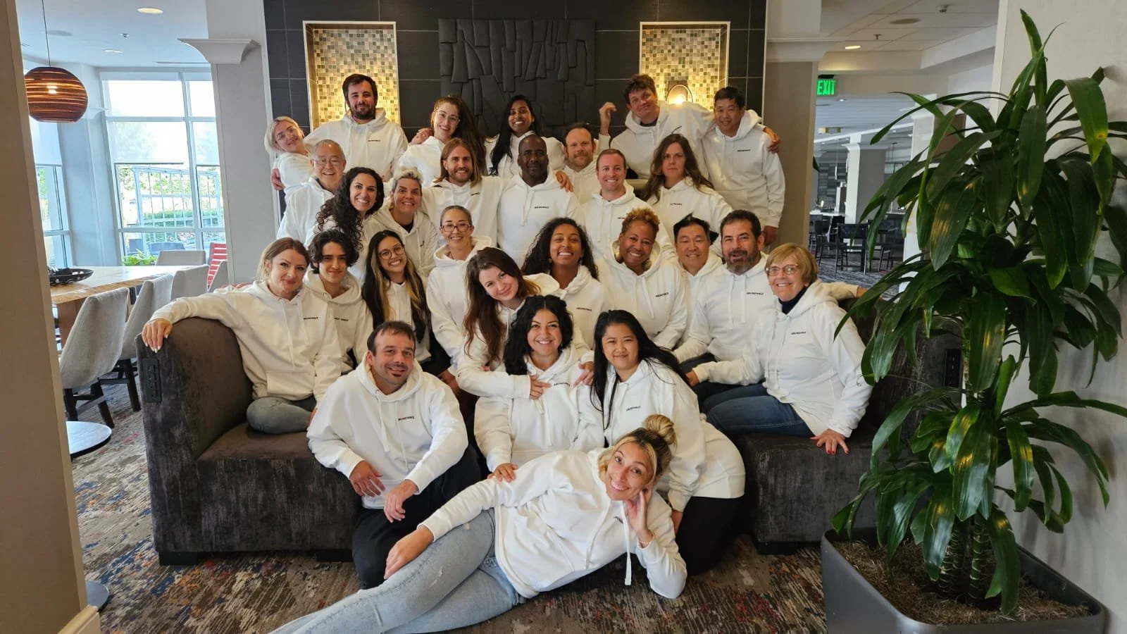 Group of people wearing white hoodies in a restaurant or lounge with modern decor and large windows. Some are sitting on a couch, others standing behind, posing for a group photo.