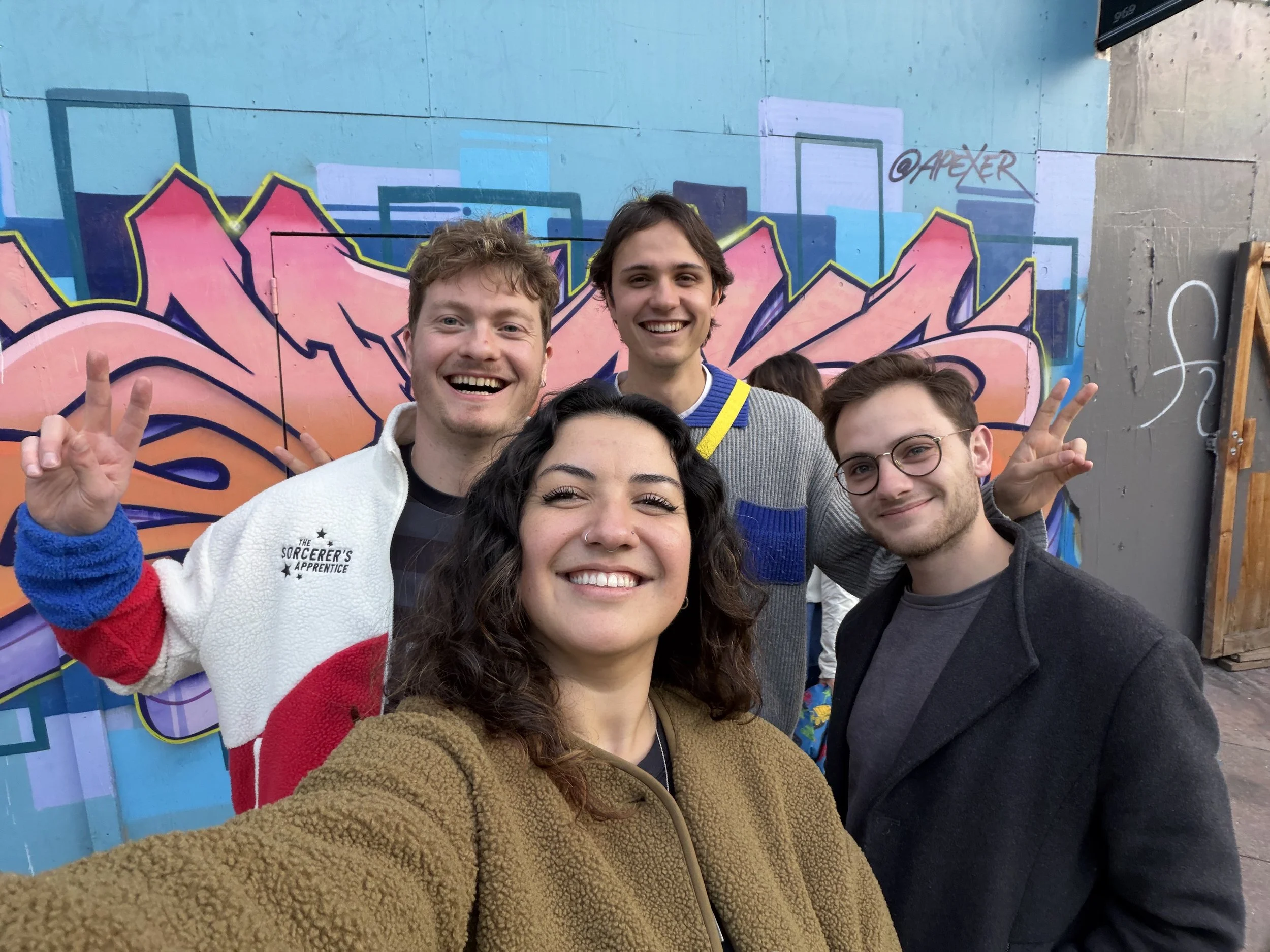A group of smiling young adults taking a selfie in front of a colorful graffiti wall. They are making peace signs and appear happy.