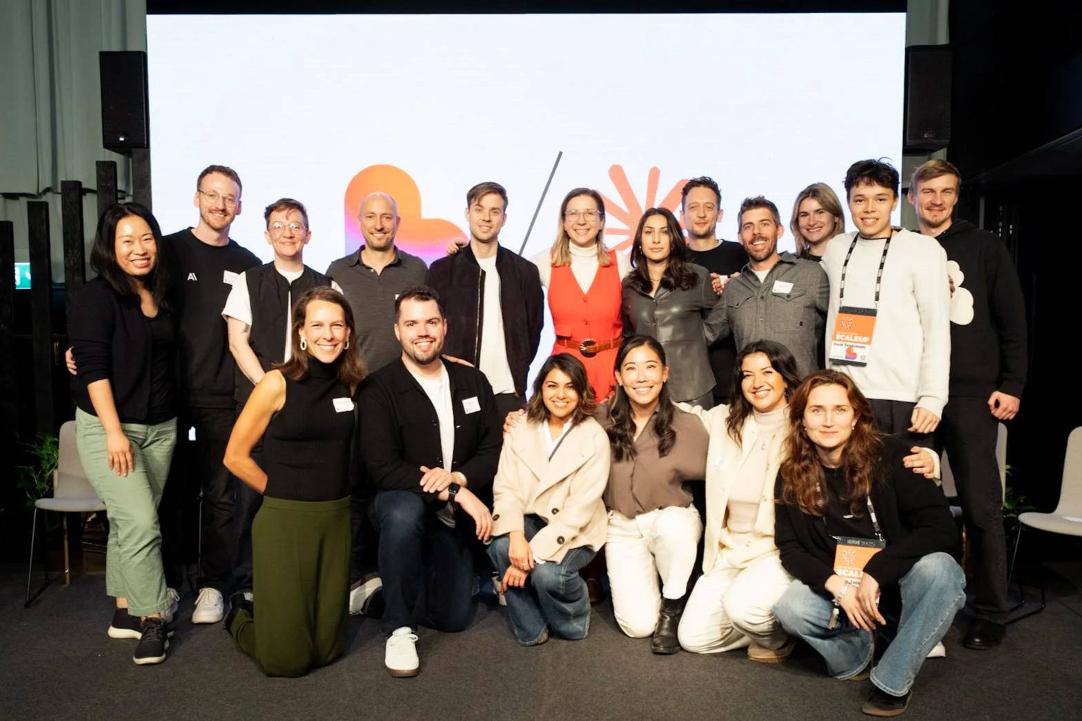 Group of people smiling and posing for a photo indoors, with a bright screen in the background displaying a logo or design.