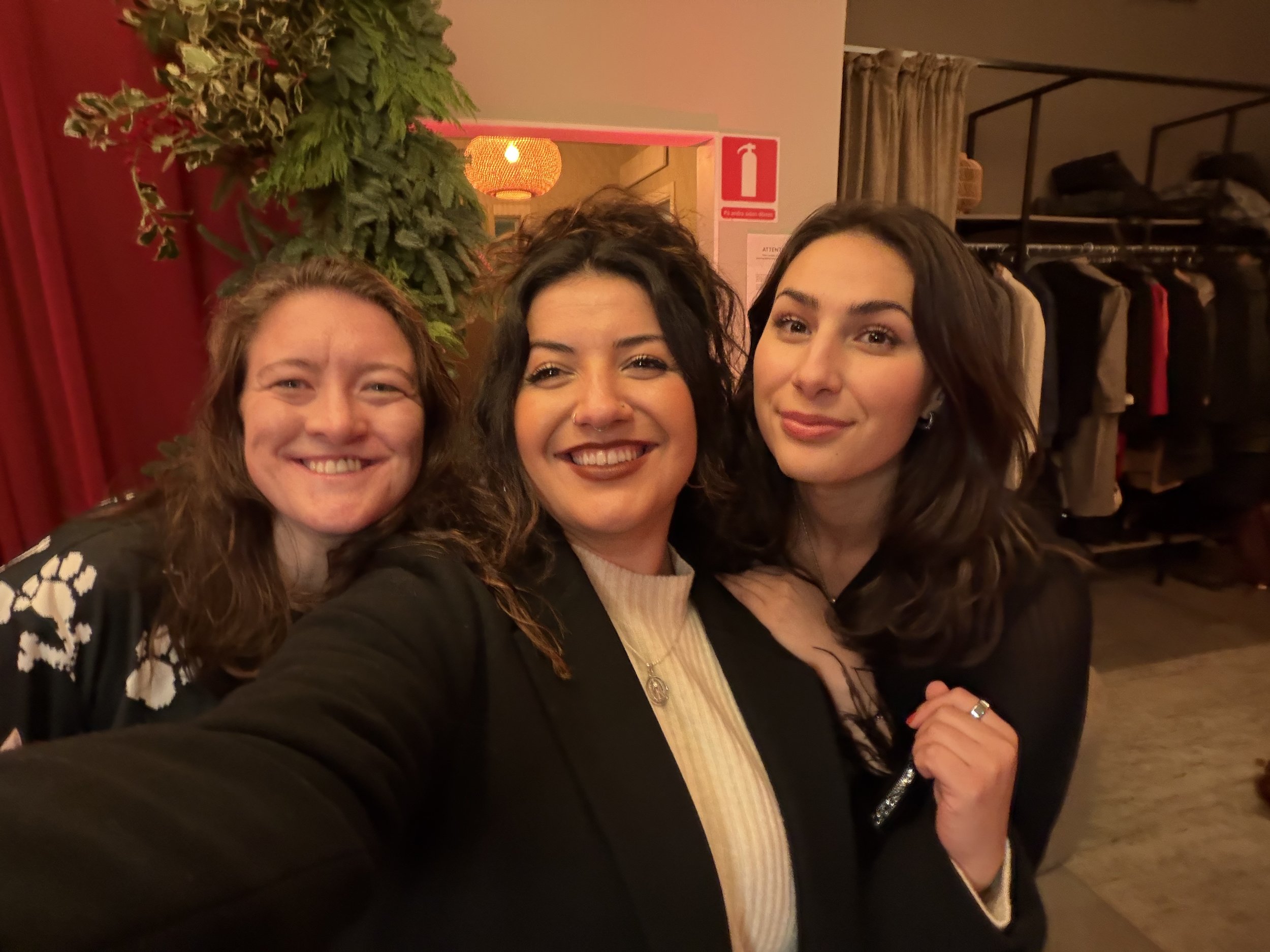 Three women smiling and taking a selfie together indoors, with a clothing rack and a curtain in the background.
