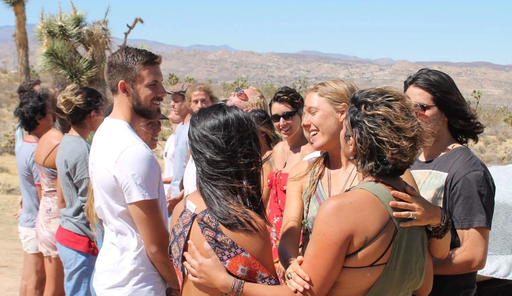 A group of people outdoors in a desert landscape, smiling and embracing each other while standing in a line.