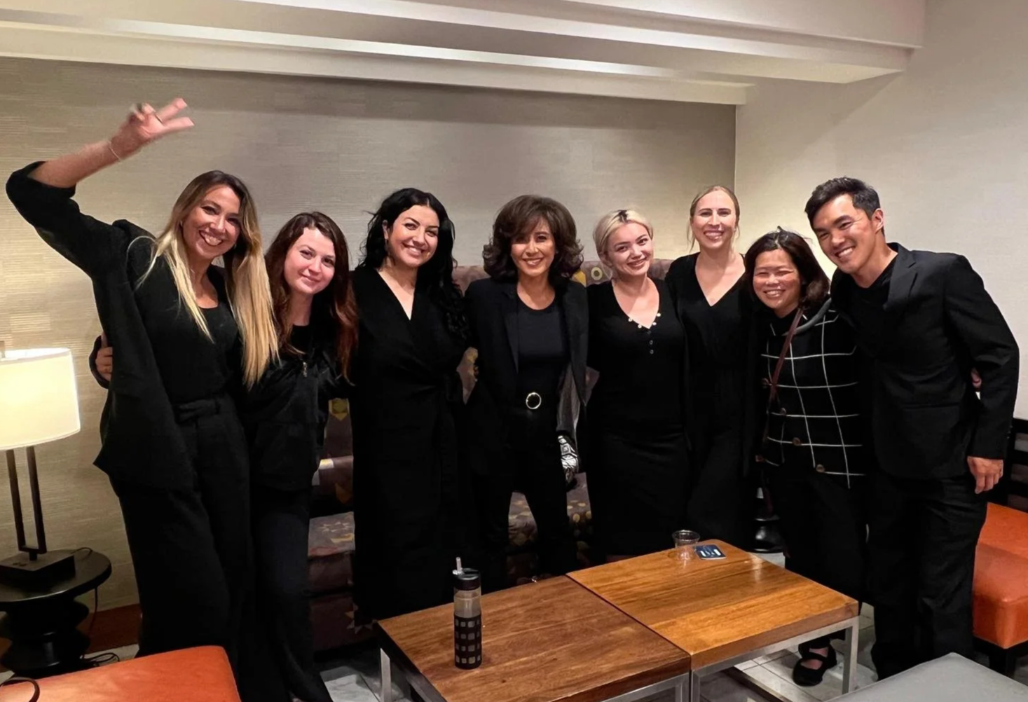 Group of nine women and one man dressed in black, smiling and standing together in a cozy indoor setting.