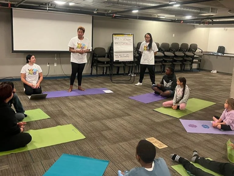 Children sitting on yoga mats in a circle during a class, with two instructors standing at the front in a room with chairs and a whiteboard.