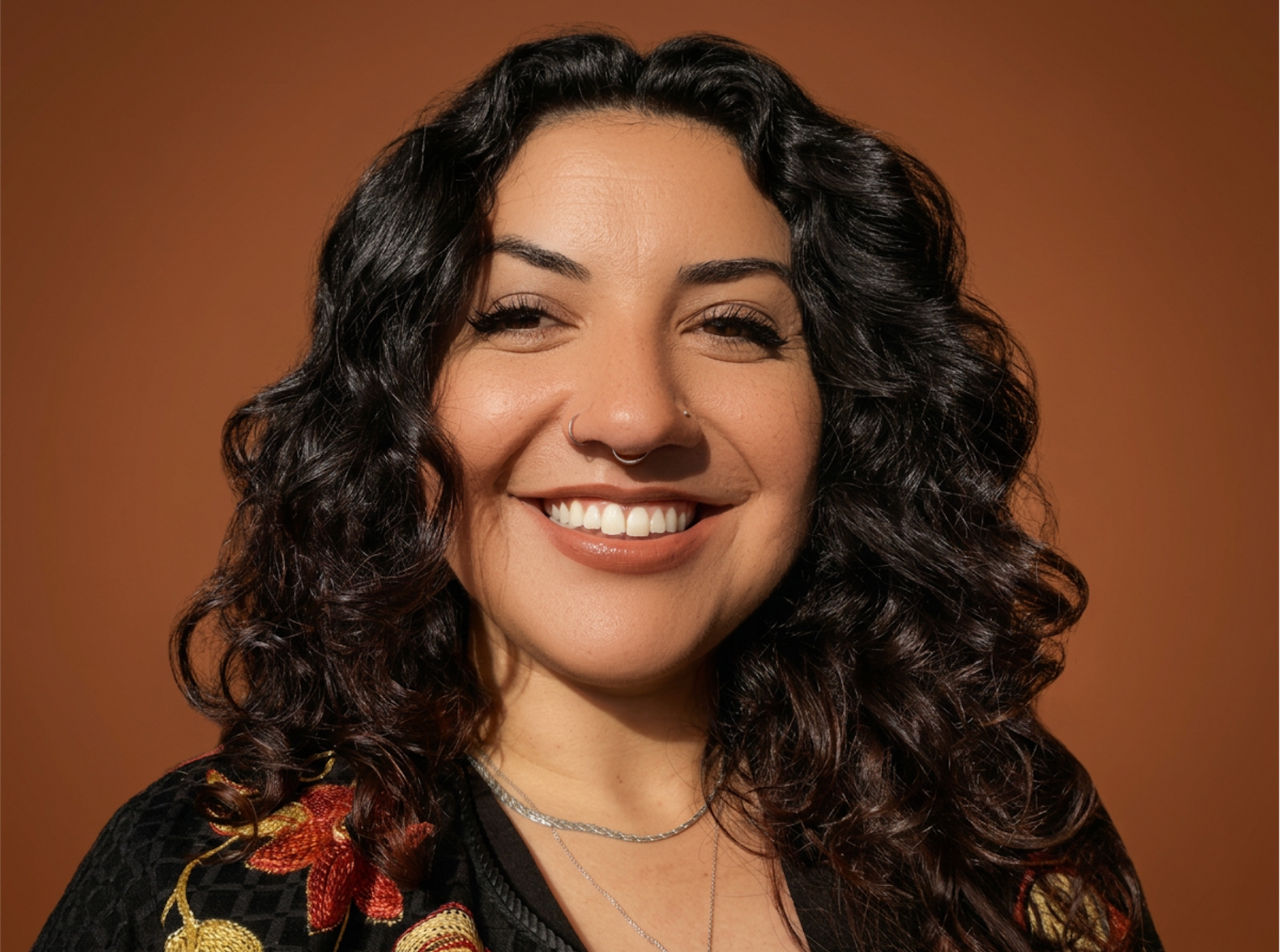 Close-up of a smiling woman with curly black hair, wearing a black top with floral embroidery, silver necklaces, and nose piercings, against a brown background.