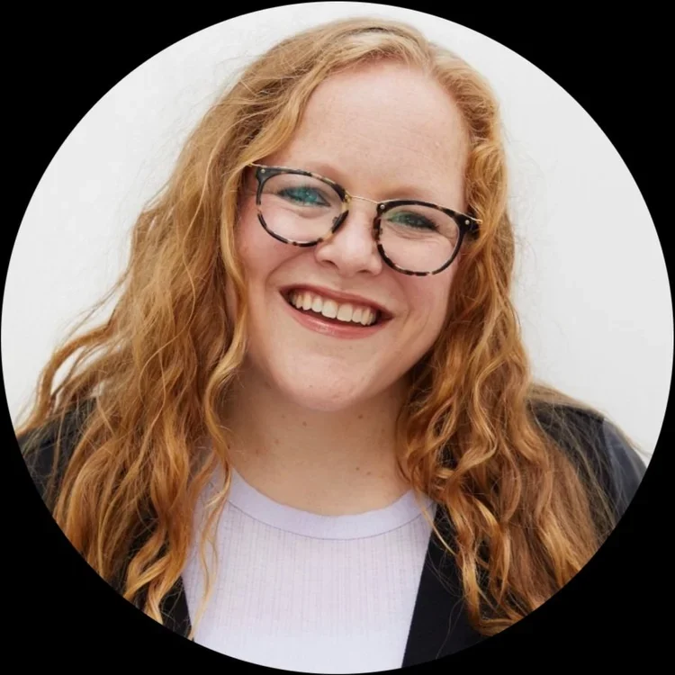 A young woman with long, curly red hair and glasses smiling at the camera against a white background.