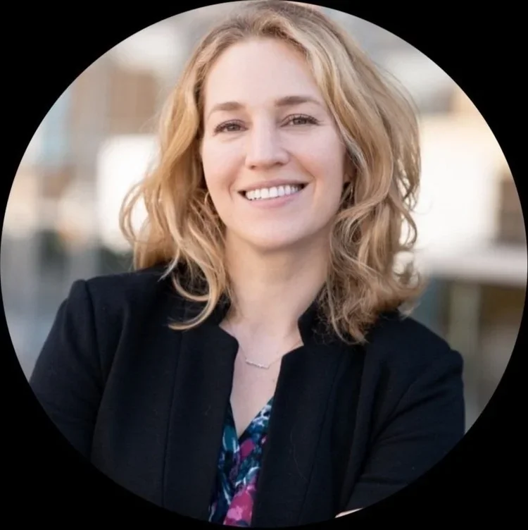 A woman with wavy blonde hair smiling, wearing a black blazer and a patterned top, outdoors with a blurred cityscape background.
