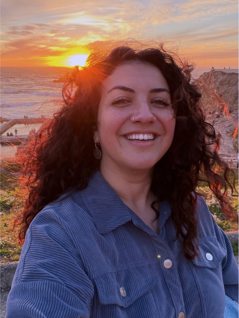 A woman with curly dark hair and earrings smiling at sunset by the ocean with a rocky coastline in the background.