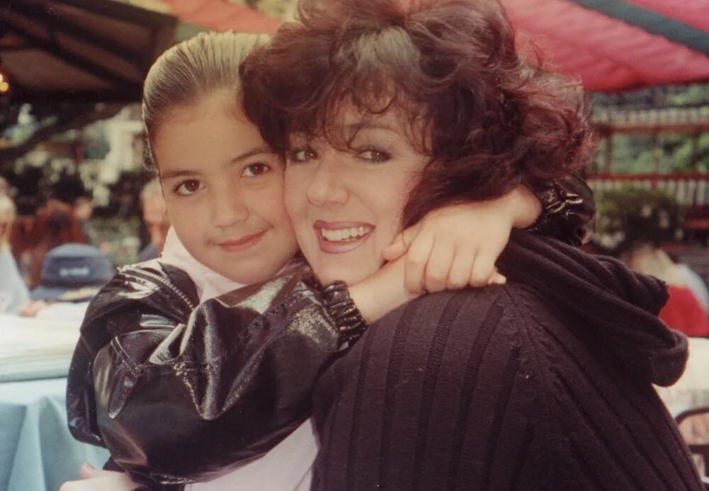 A young girl with brown hair and a woman with curly brown hair hugging each other and smiling at the camera in an outdoor setting.
