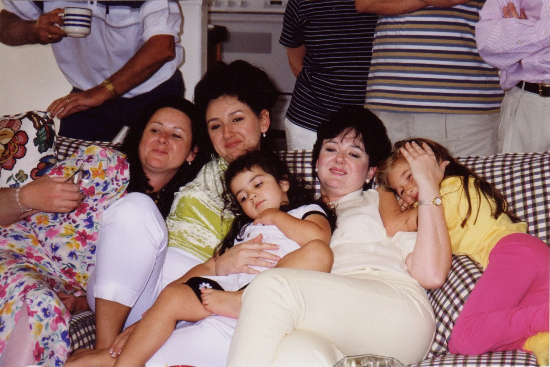 A group of women and children lying on a couch, relaxing and smiling. Some stand behind them, partially visible, in casual attire.