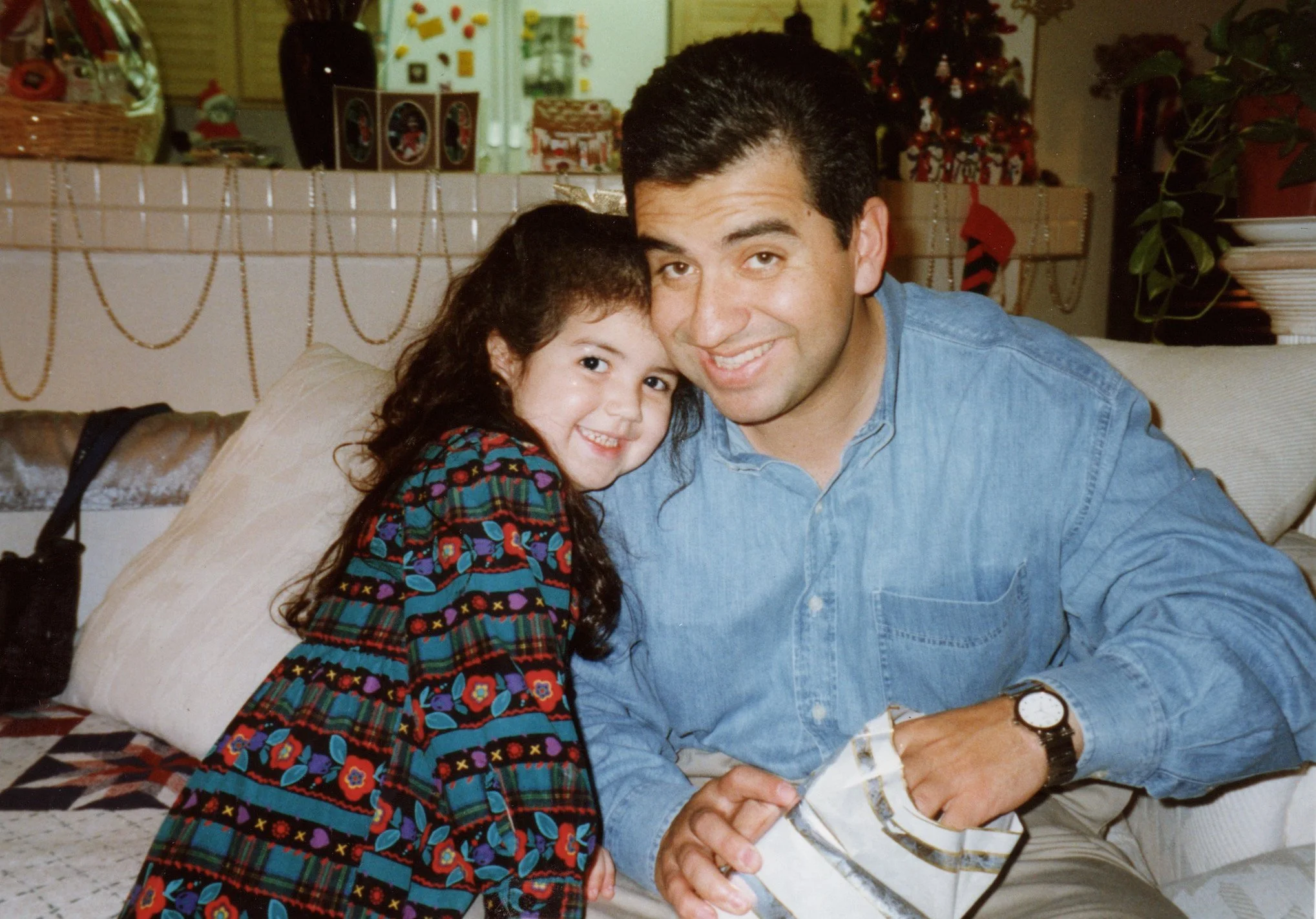 A man and a young girl sitting together on a couch, smiling and holding hands. The man is wearing a blue denim shirt and a watch, and the girl is dressed in a colorful, patterned dress. In the background, there are Christmas decorations including a decorated tree, stockings, and holiday-themed items.
