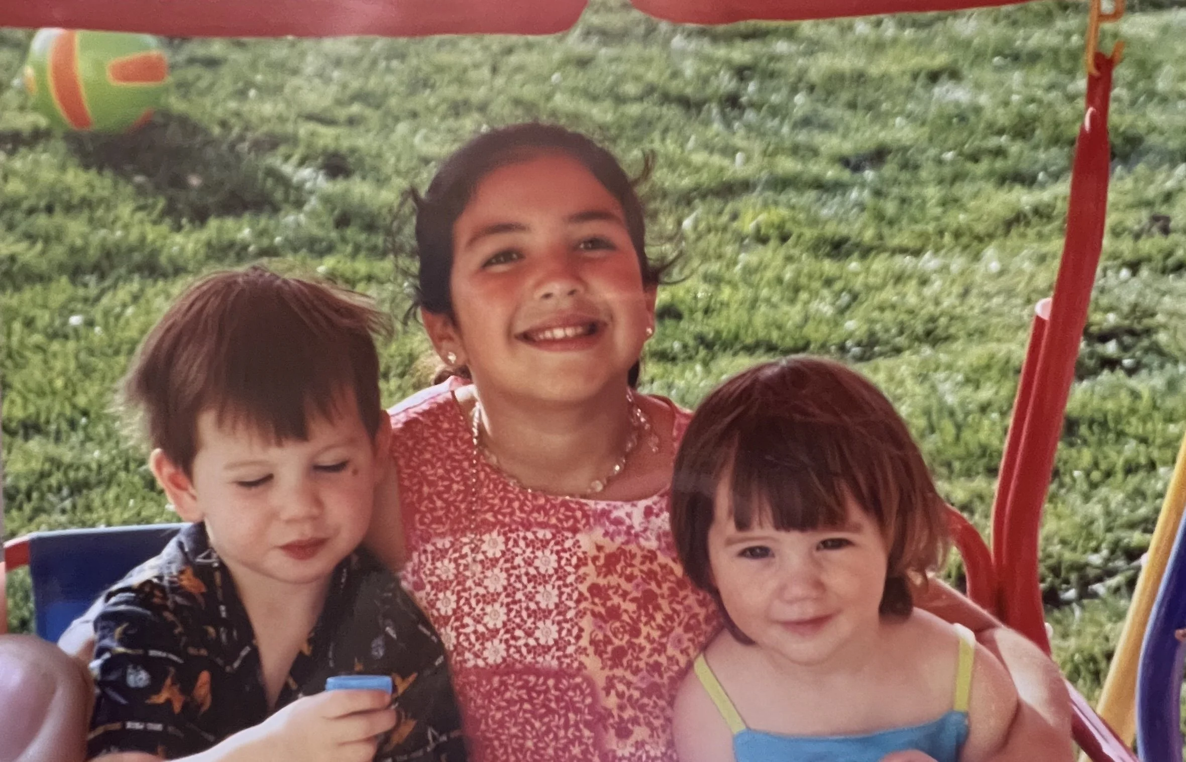 Four children sitting close together outdoors, with a grassy background and part of a playground structure visible. One girl in the center is smiling, wearing a red dress with white patterns, and has earrings. Two boys and another girl are also visible.