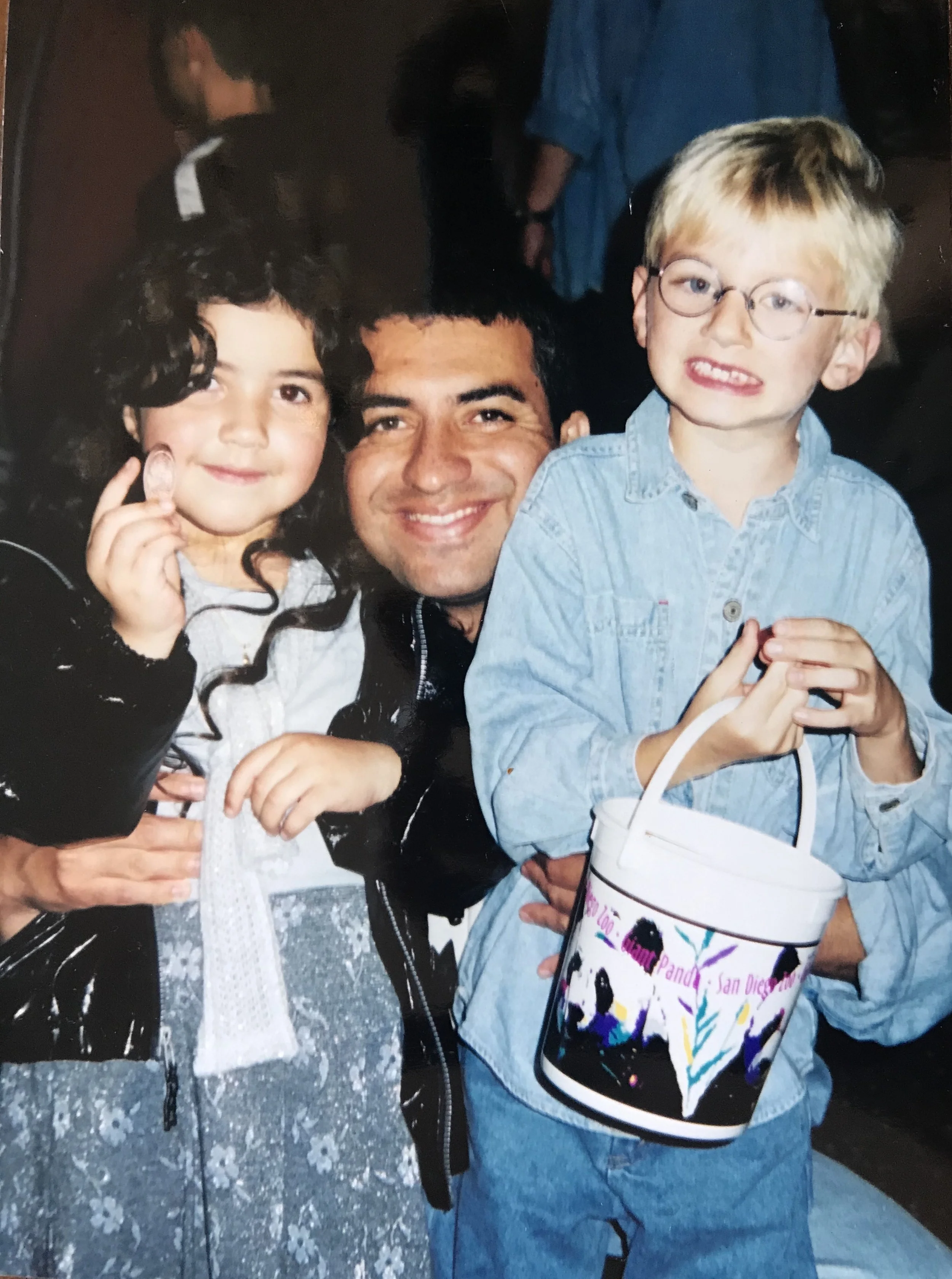 A group of three people, two children and one adult, smiling and posing for a photo inside a festive or event setting. The girl on the left has dark, curly hair and is holding an ice cream cone. The boy on the right has blonde hair, glasses, and is holding a colorful bag with illustrations of women and the text 'San Diego' visible. The adult in the middle has dark hair and is smiling. There is a bucket or container held by the boy with a strap, possibly a backpack or gift bag, and the background features dark, indistinct figures.