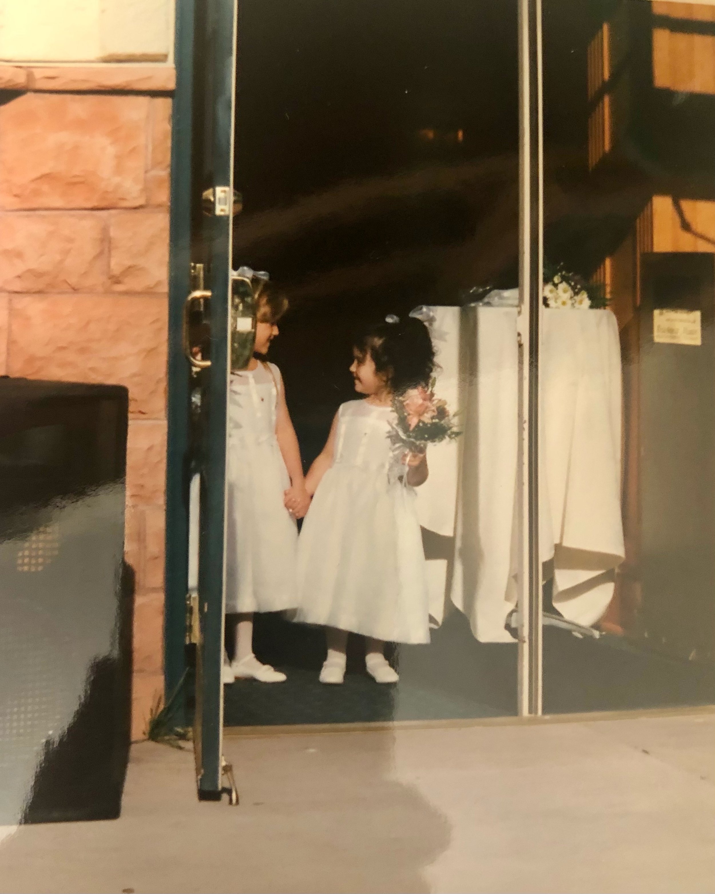 Reflection of two women in the mirror, one with curly dark hair and the other with short light brown hair, holding hands, wearing white dresses, and standing in a room with brick wall and wooden furniture.