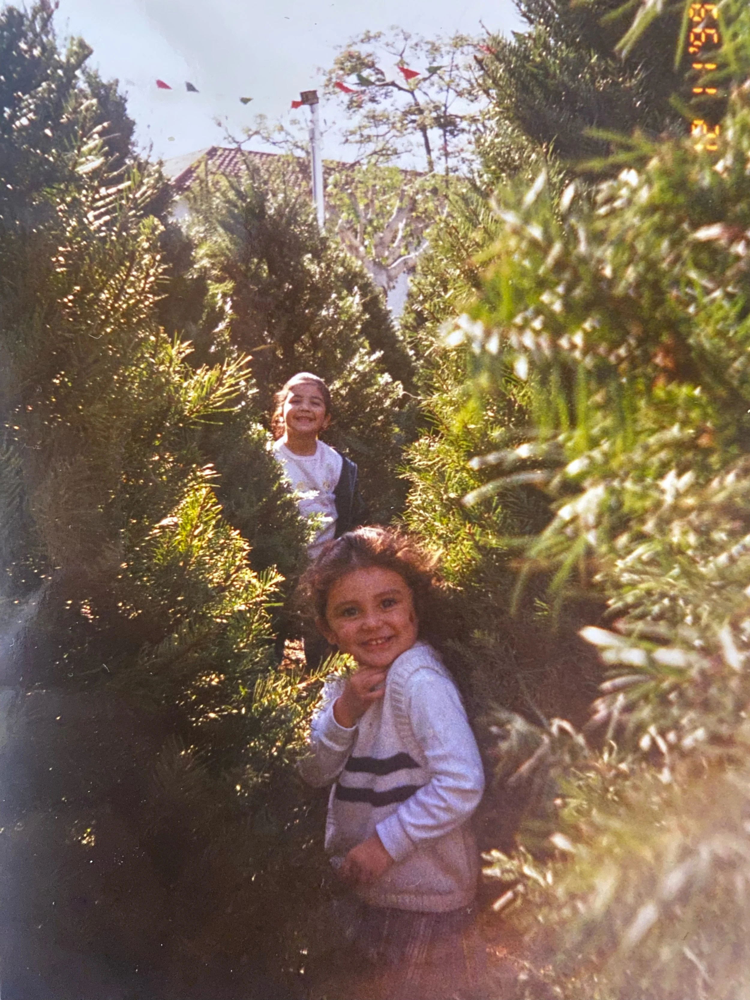 Two children with light skin standing among dense green bushes, one girl smiling and wearing a white top with black stripes, another girl in the foreground with a smile, wearing a white sweater, in an outdoor setting with trees and a house with a tiled roof in the background.