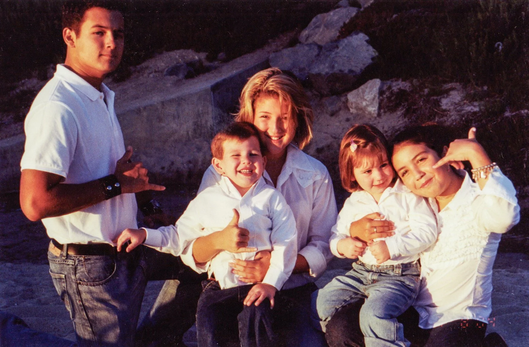 A group of five children and one adult woman posing outdoors near rocks at sunset, with the children smiling and making gestures, dressed in white shirts.