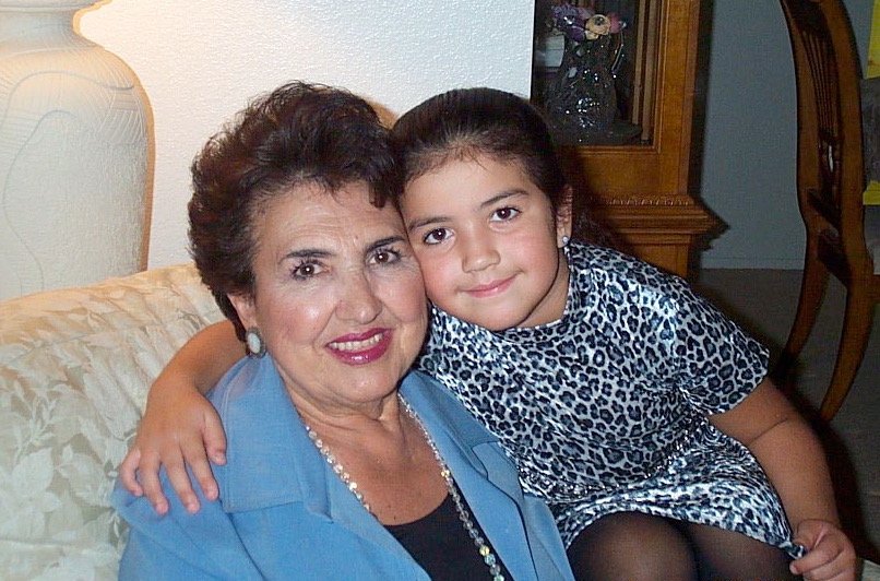 A woman and a young girl sitting on a couch, hugging and smiling at the camera, in a living room with a wooden cabinet in the background.