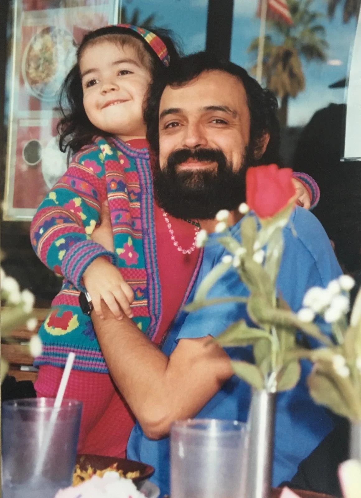 A man with a beard and a blue shirt is holding a young girl with curly hair and a colorful sweater. They are smiling at the camera with a blurred restaurant background, a flower in the foreground, and drinks on the table.