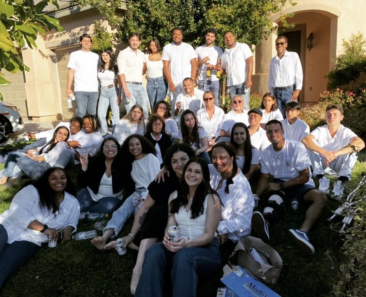A large group of people gathered outside a house, posing for a photo, some sitting on the grass, others standing, with a sunny backdrop and greenery around.