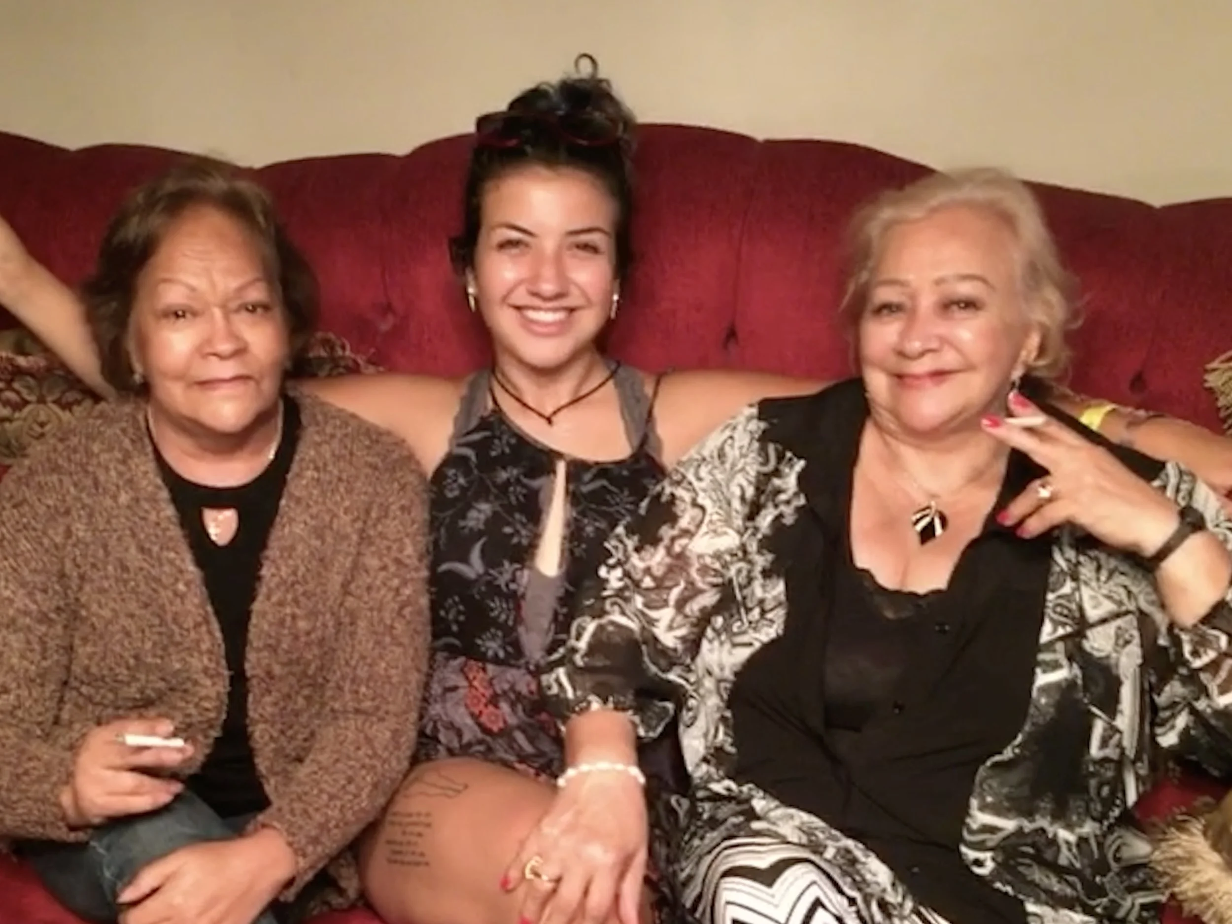 Three women sitting close together on a red sofa, smiling at the camera. The woman in the middle has dark hair, the woman on the left has short brown hair, and the woman on the right has blond hair. They are wearing casual clothing and are in a cozy indoor setting.