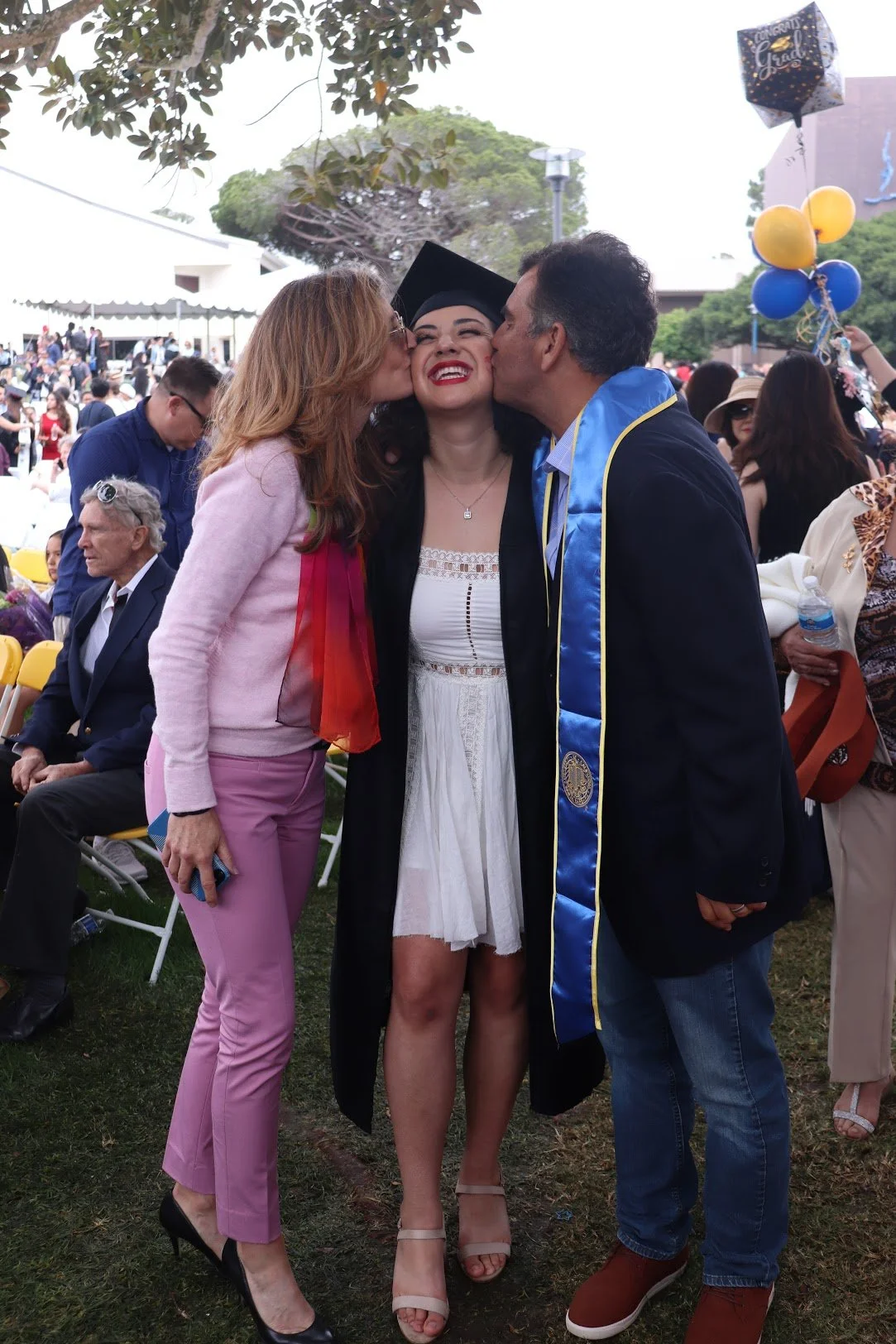A graduating woman in a gown and cap receives kisses on each cheek from two people at an outdoor graduation ceremony with many seated and standing attendees, balloons, and trees in the background.