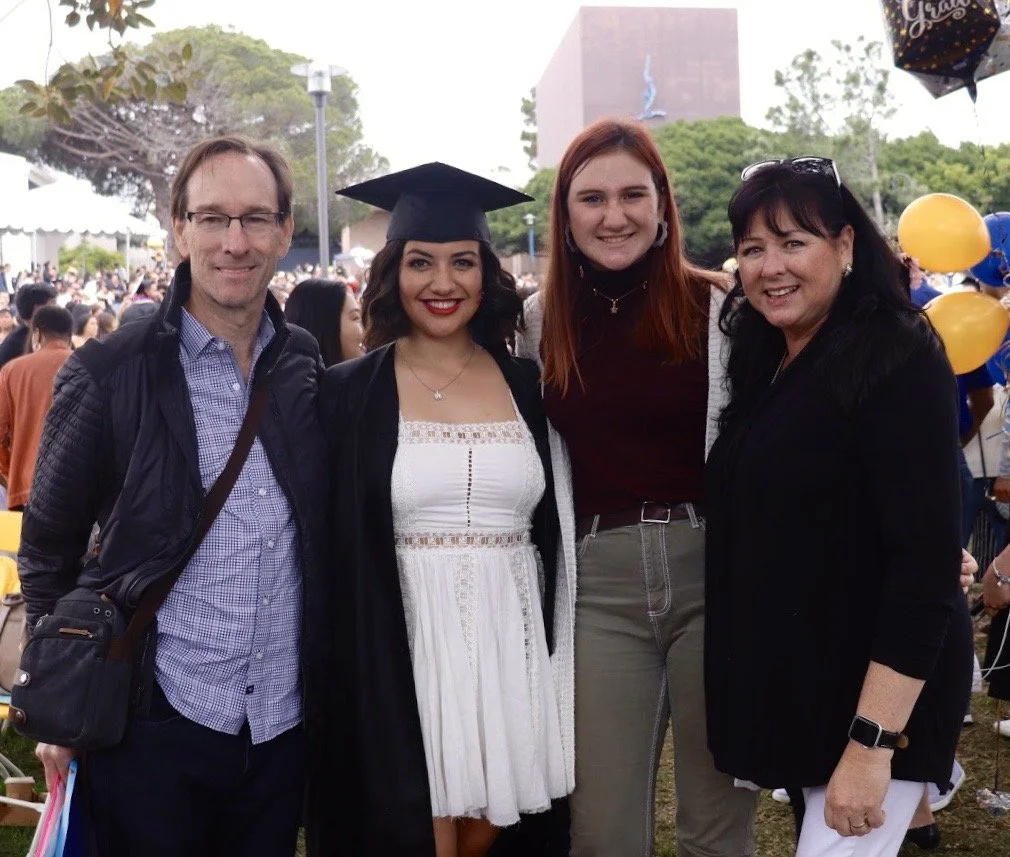 A group of four people at a graduation celebration outdoors, one woman wearing a graduation cap and gown, surrounded by friends and family.