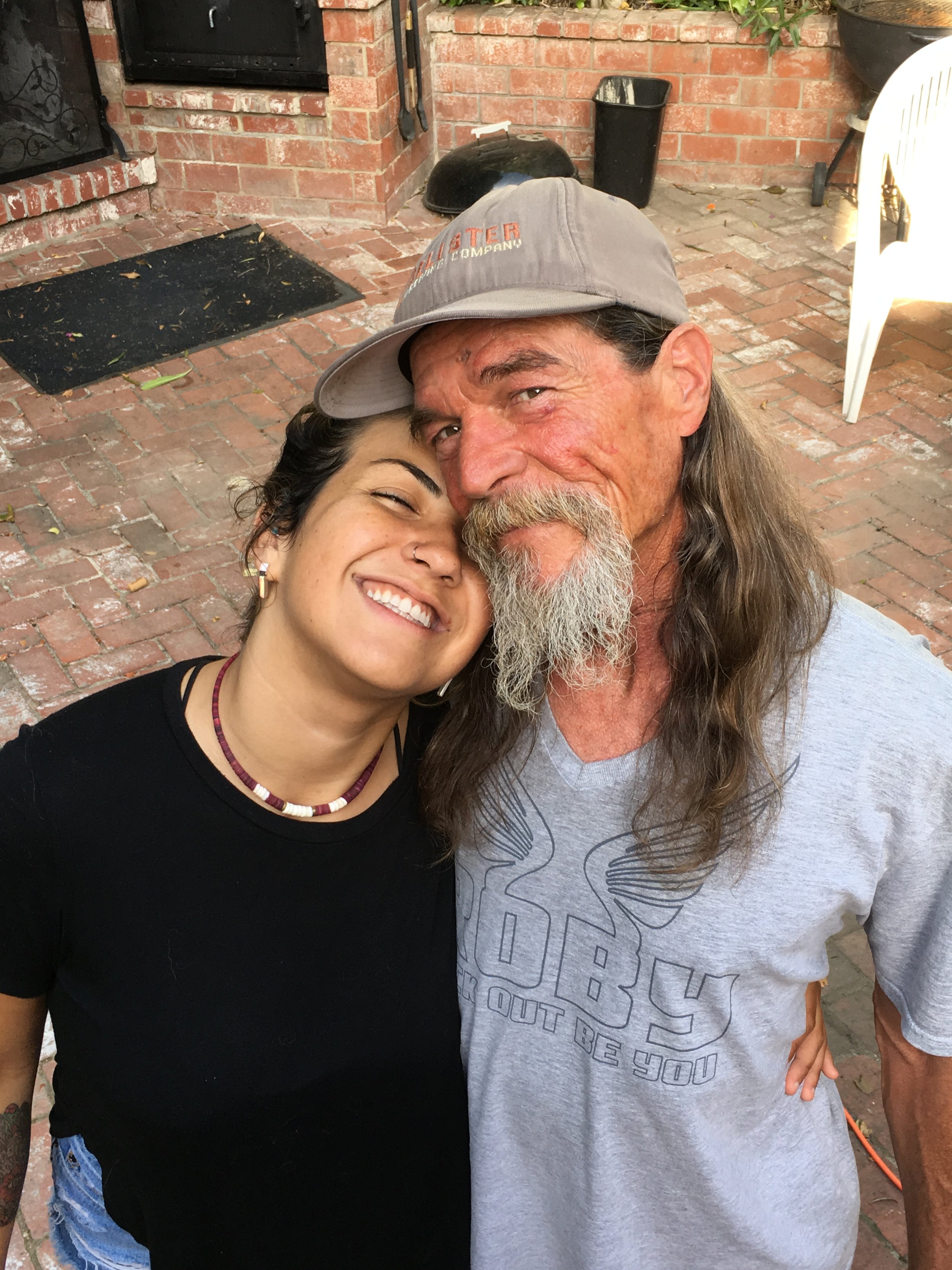 Two people, a young woman and an older man with long hair and a beard, are taking a selfie together outdoors on a brick patio. The woman is smiling with her eyes closed, and both are leaning into each other. The background includes a brick fireplace with a black door, a trash can, a black grill cover, and some patio furniture.