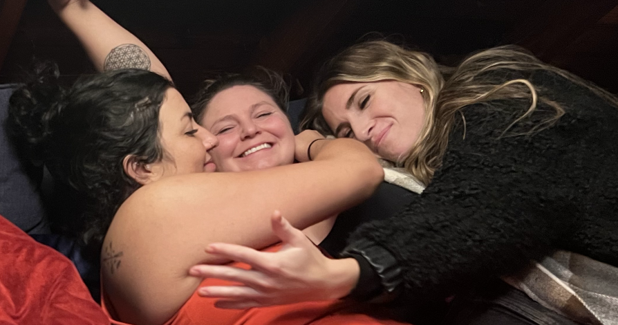 Three women cuddling and smiling on a dark couch, embracing each other affectionately.