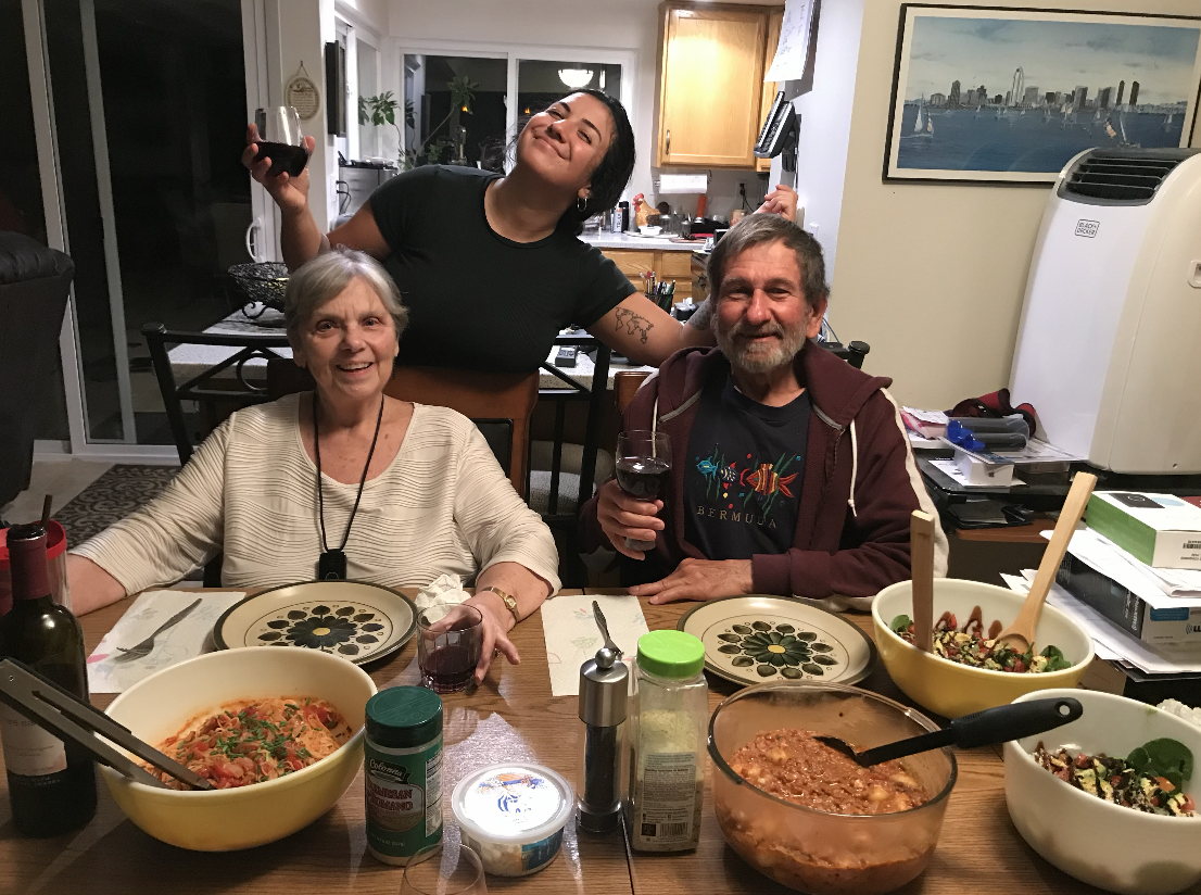 A group of three people celebrating at a dinner table with various dishes and drinks, smiling and holding glasses of wine, in a cozy home setting.