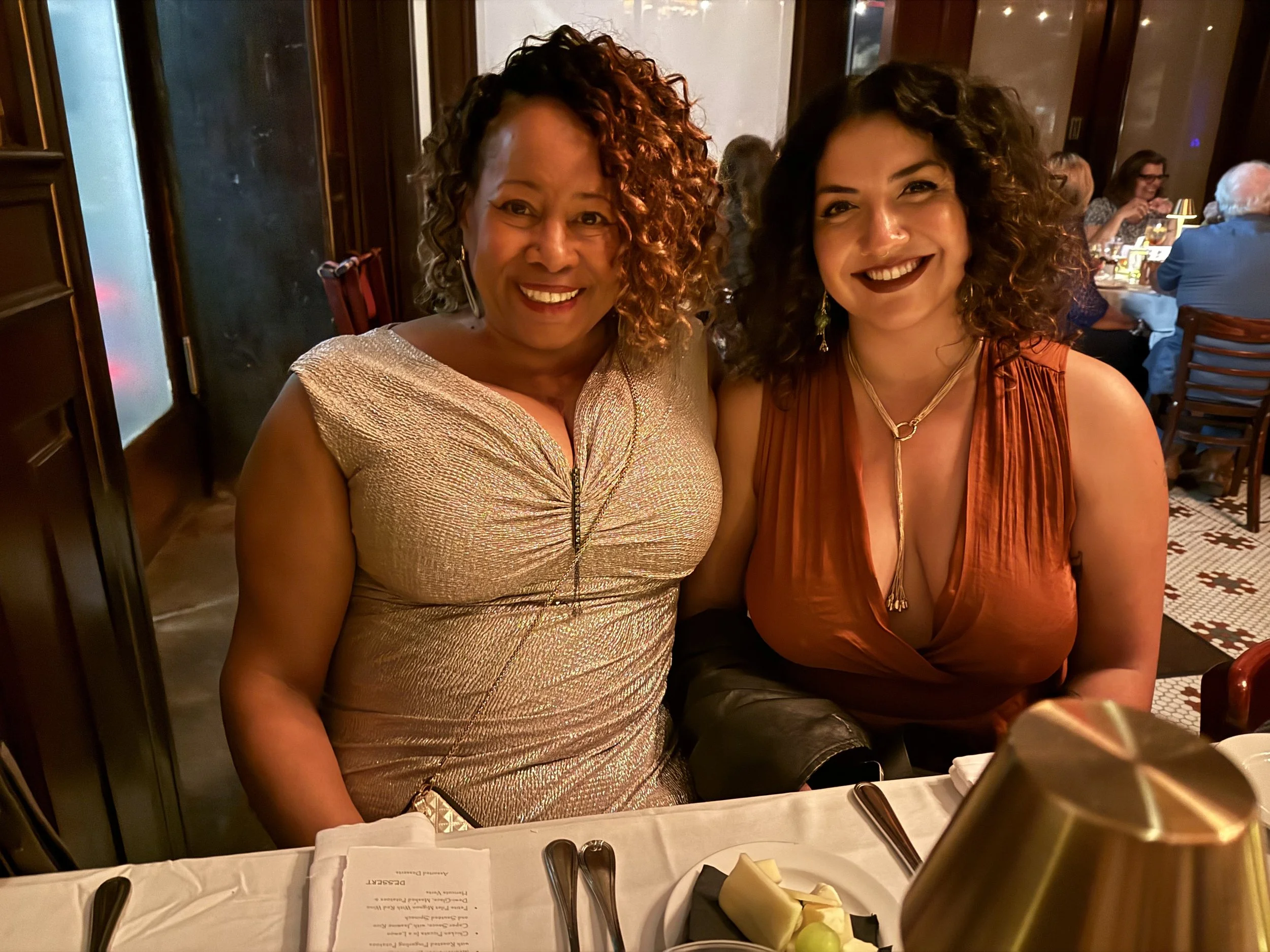 Two women smiling and sitting together at a dinner table in a restaurant, dressed in elegant clothing, with a dinner menu, utensils, and a small decorative plant on the table.