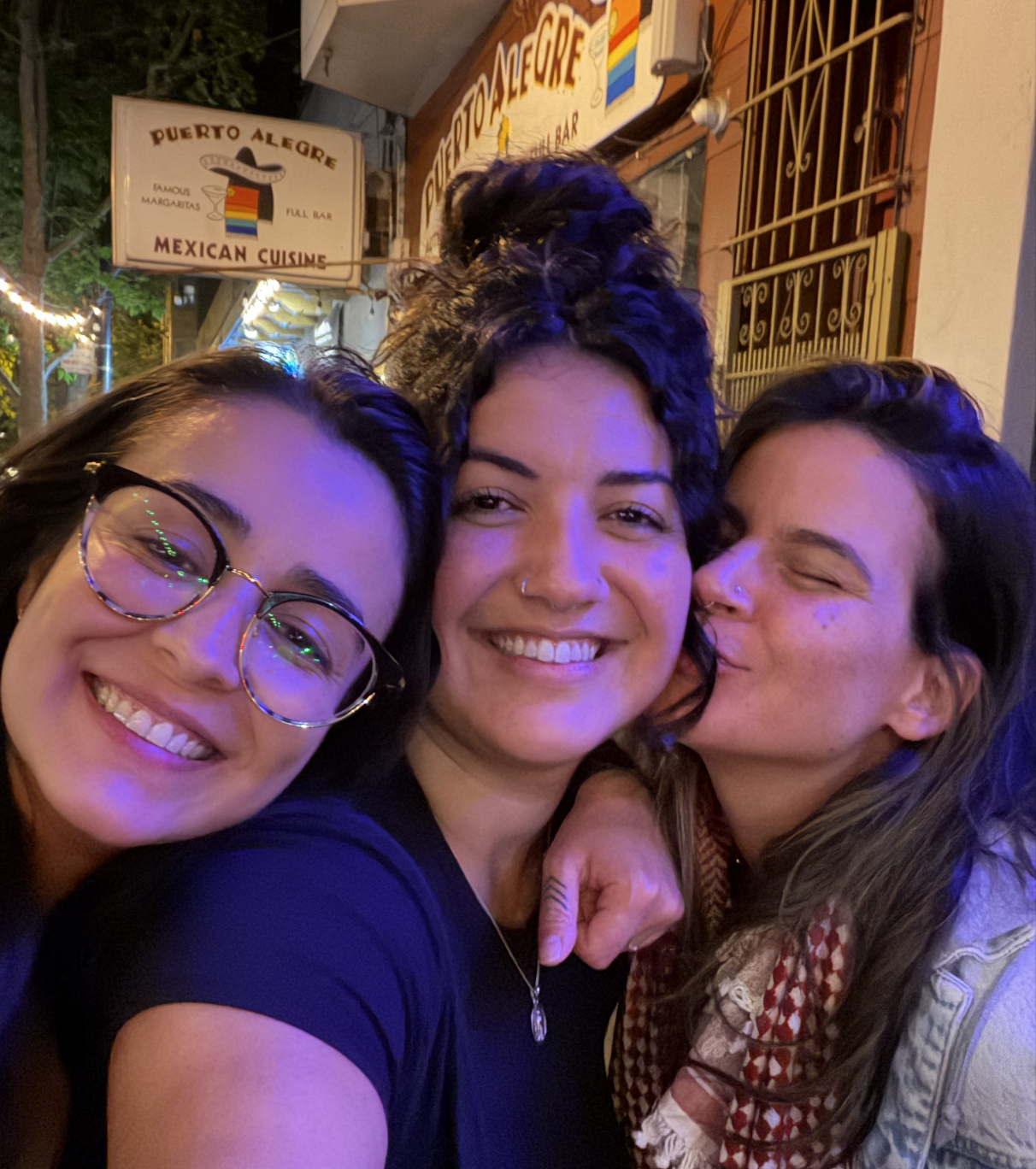 Three women taking a selfie outside a Mexican restaurant at night. They are smiling and appearing happy, with one woman kissing another on the cheek. The background shows signs for "Puerto Alegre" and "Mexico Cuisine."