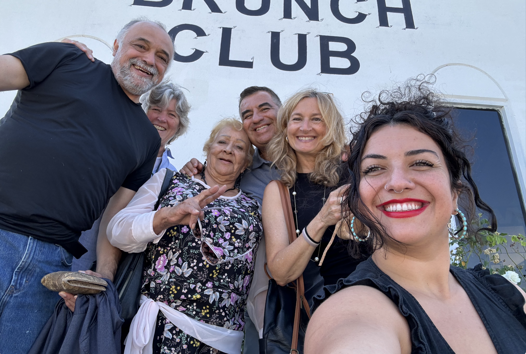 A group of seven smiling people, including three older women and four younger adults, posing for a selfie outside a building with a sign that says 'DUNEDIN CLUB.'