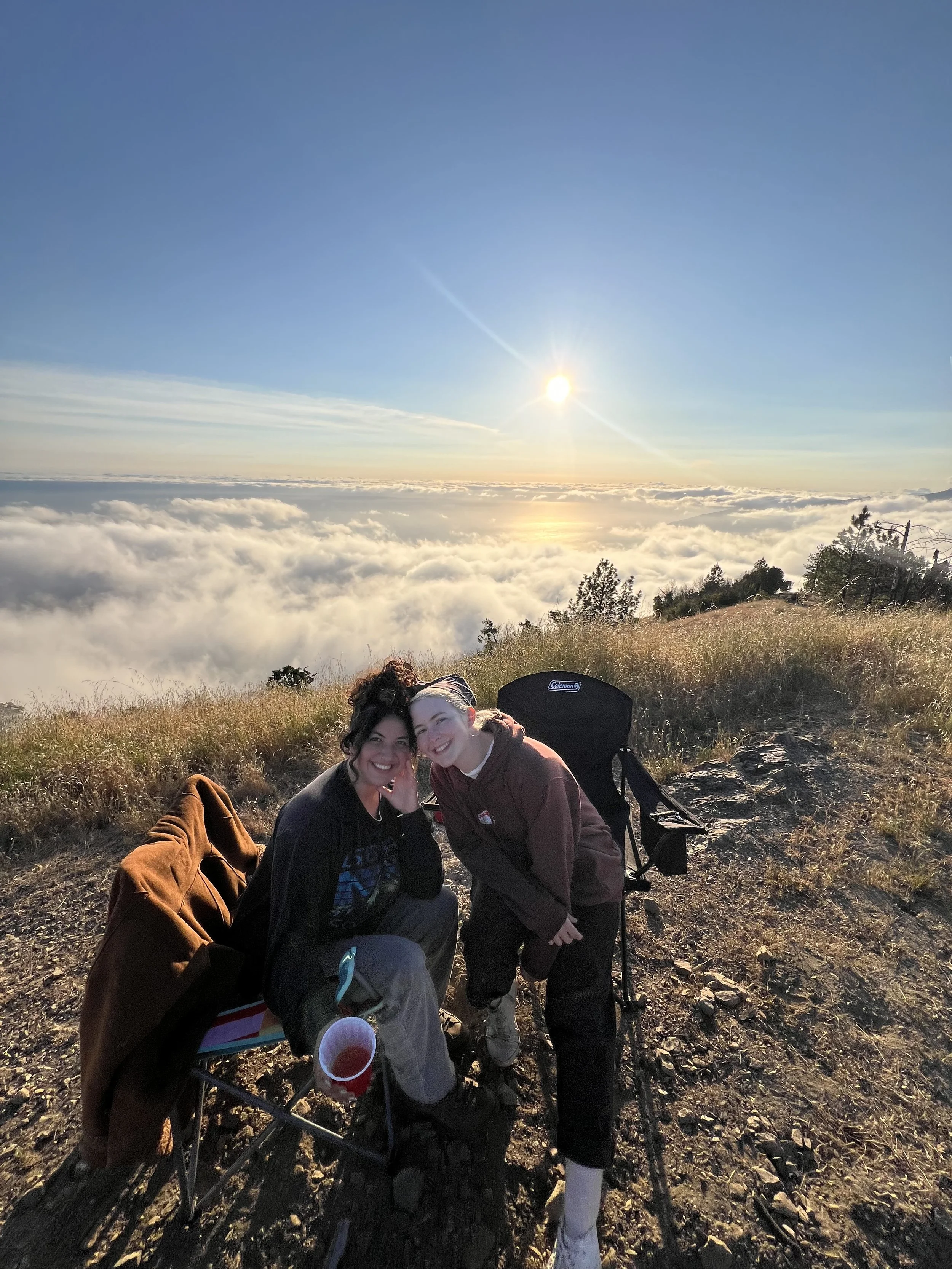Two smiling women sitting on the ground on a hillside at sunset, with a view of clouds and the sun low in the sky behind them. One woman has darker hair and the other has lighter hair. They are surrounded by a backpack and a chair.