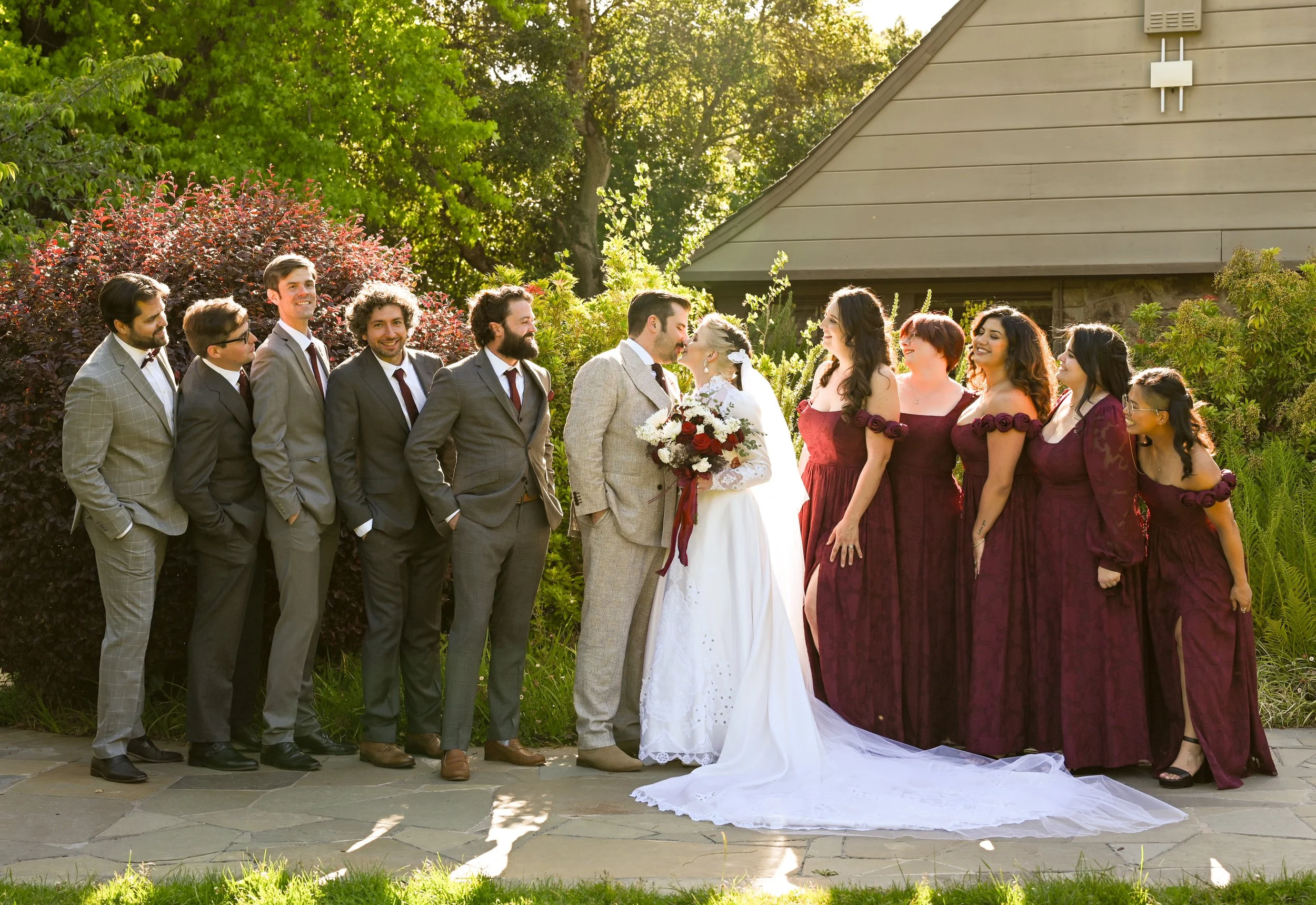 Group of wedding guests standing outdoors, with a bride and groom kissing in the center, surrounded by greenery and sunlight.