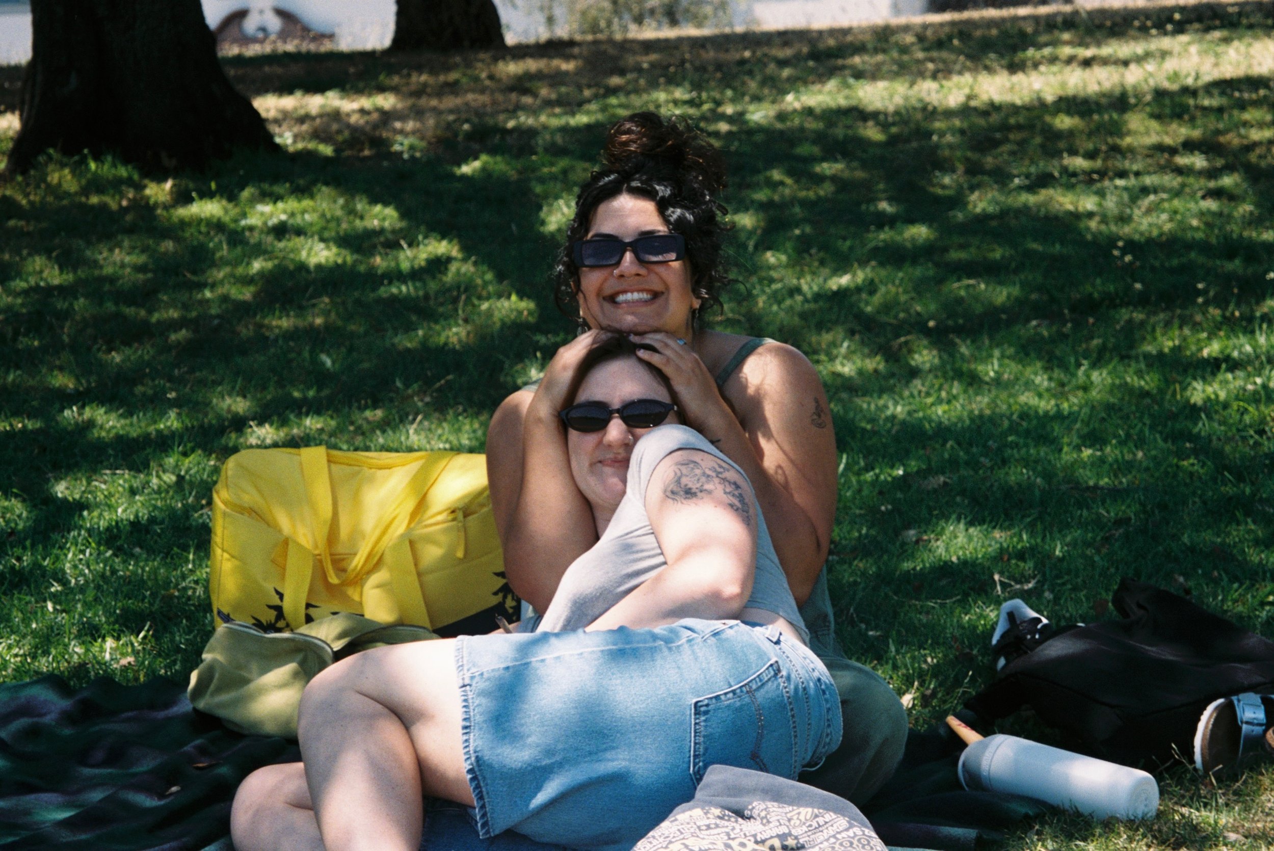 Two women lying on the grass in a park, smiling and wearing sunglasses, with a tree and a white building in the background.