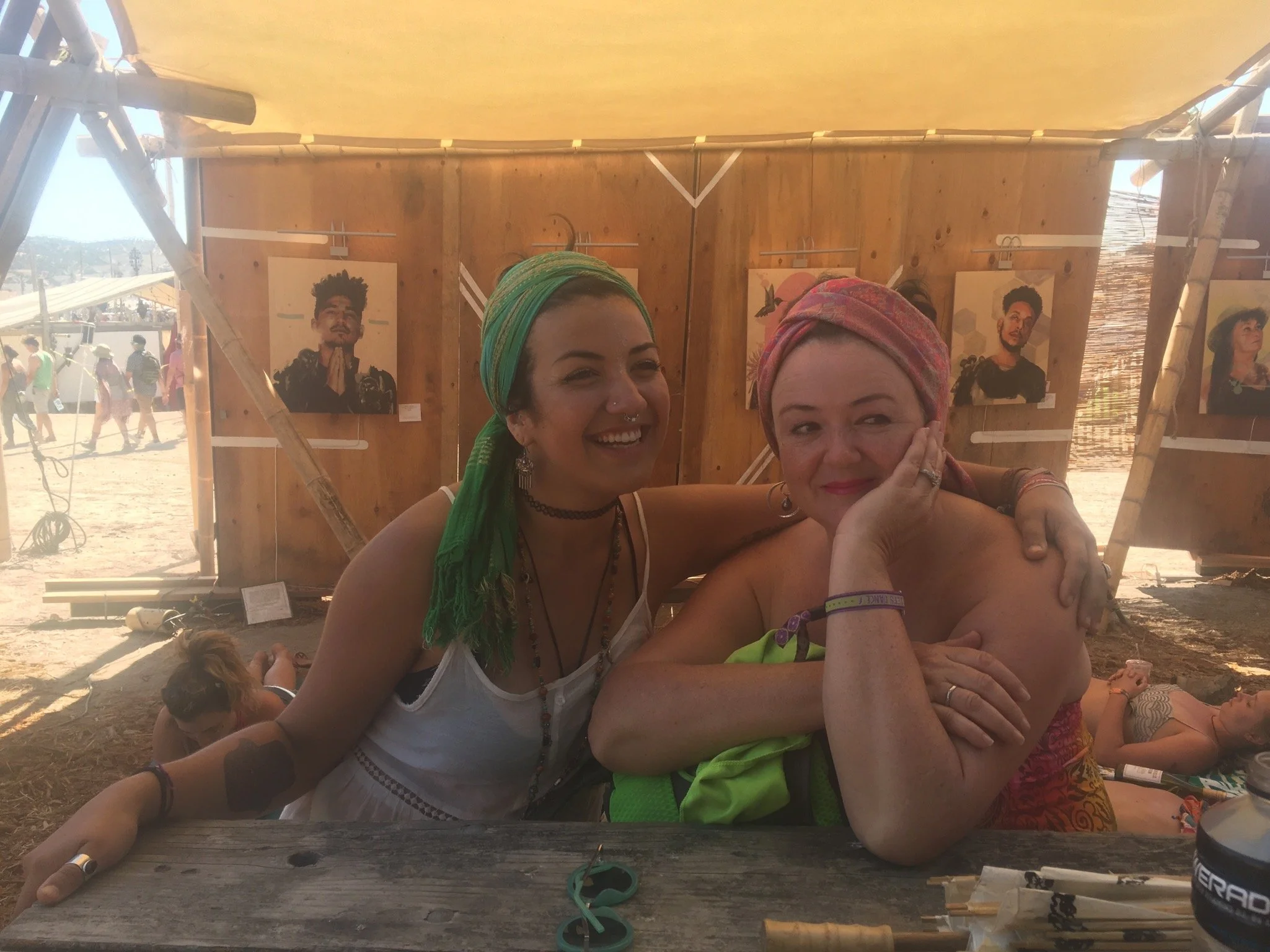 Two smiling women with colorful head wraps sitting at a wooden table, with artwork displayed in the background at an outdoor event or festival.