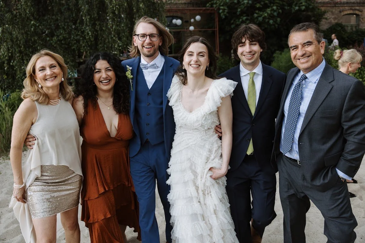 A group of six people smiling and posing outdoors at a wedding, with a bride in a white dress in the center.
