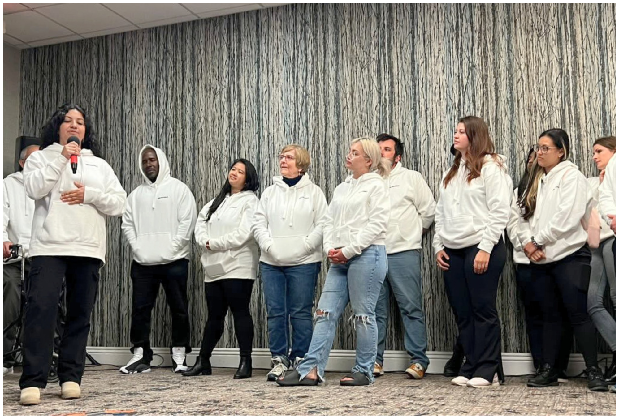 Group of diverse women standing together in a room with a textured gray wall, wearing white hoodies, some with their hands over their hearts, listening attentively to a woman speaking into a microphone.