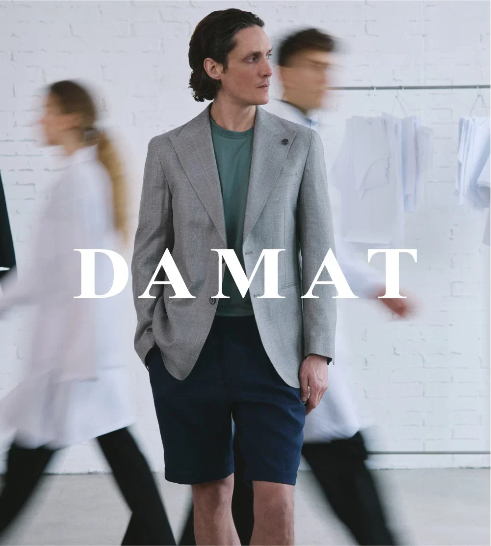Man wearing a gray blazer and shorts, standing in a room with white brick walls, with blurred people walking past.
