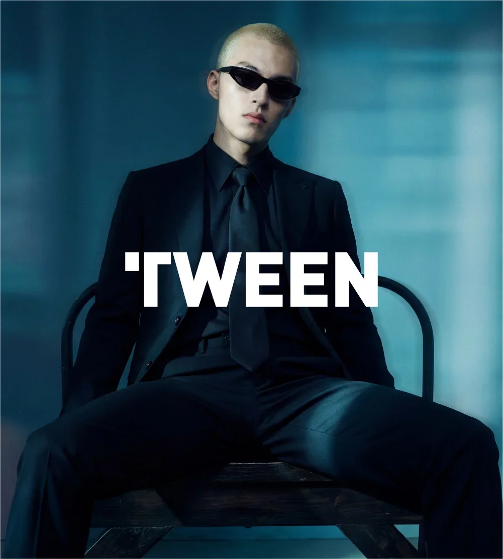 Young person with bleached hair wearing black sunglasses, black suit, and tie, sitting on a bench against a blue background with the word 'TWEEN' overlaid.