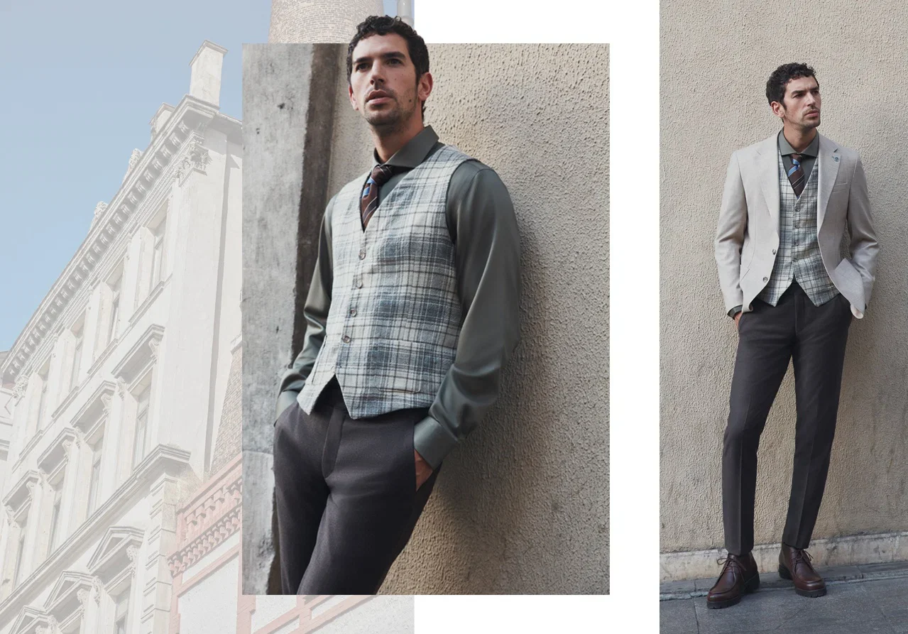 A man dressed in a stylish, layered outfit consisting of a grey blazer, plaid vest, and dark pants, standing against a textured wall in an urban setting.